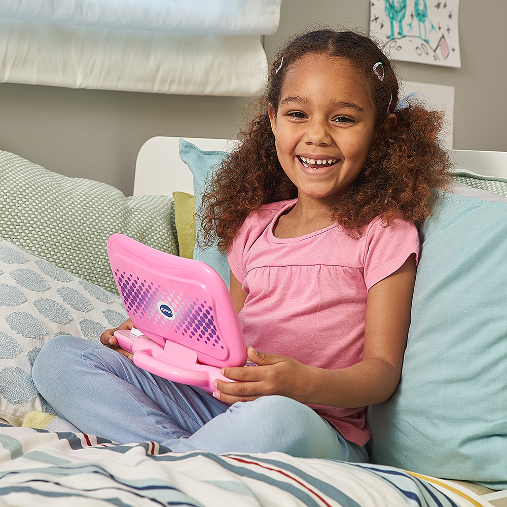 Ein Mädchen lächelt, sitzt auf einem Bett und hält einen rosa Spielzeuglaptop.