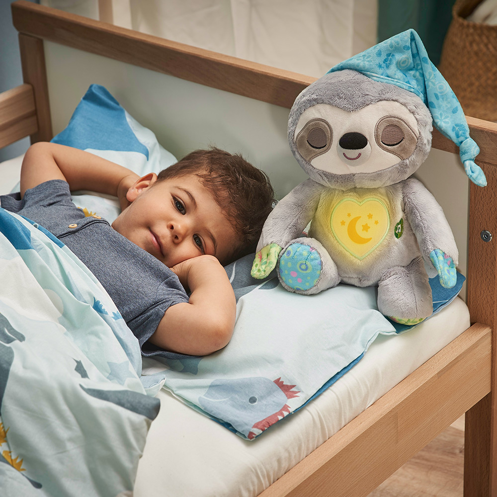 Ein Kind liegt im Bett mit einem Plüschfaultier. Das Kind schaut in die Kamera.