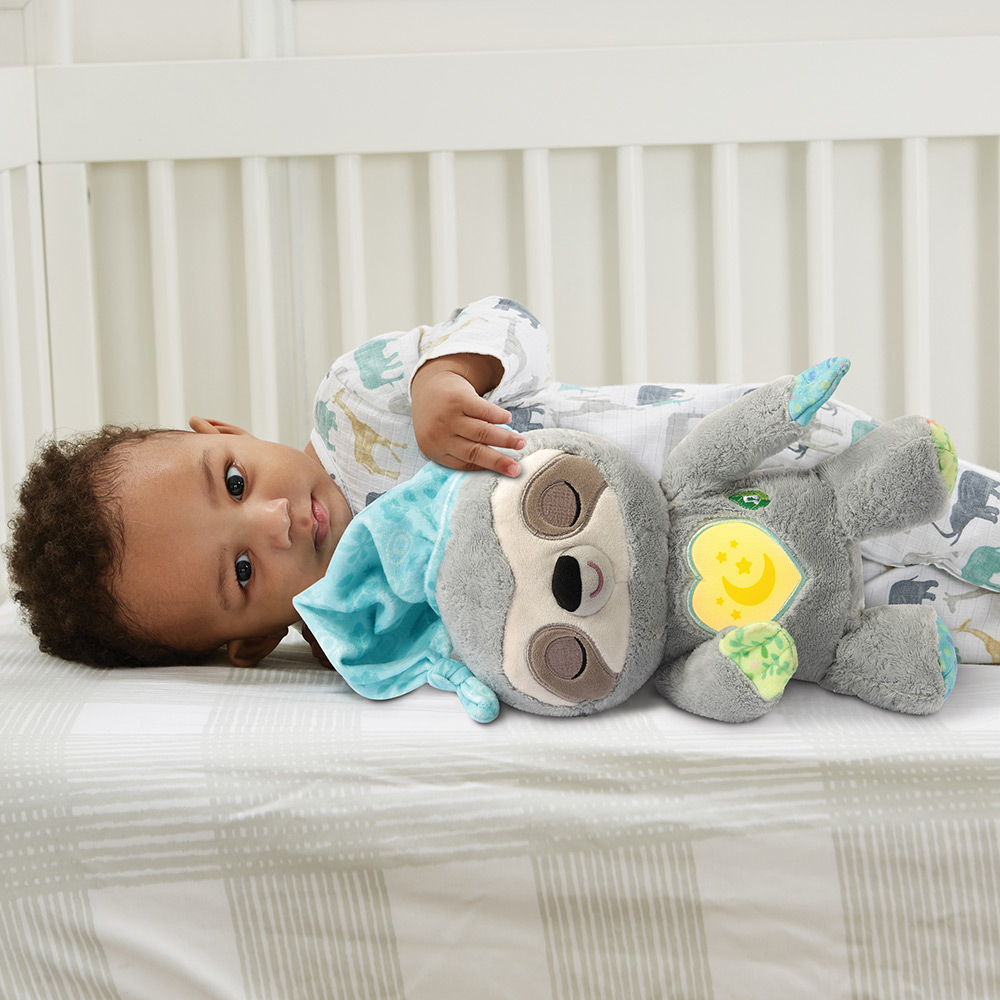 Baby mit dunklen Haaren liegt auf einem Bett mit einem Stofftier-Faultier, trägt einen gemusterten Strampler.