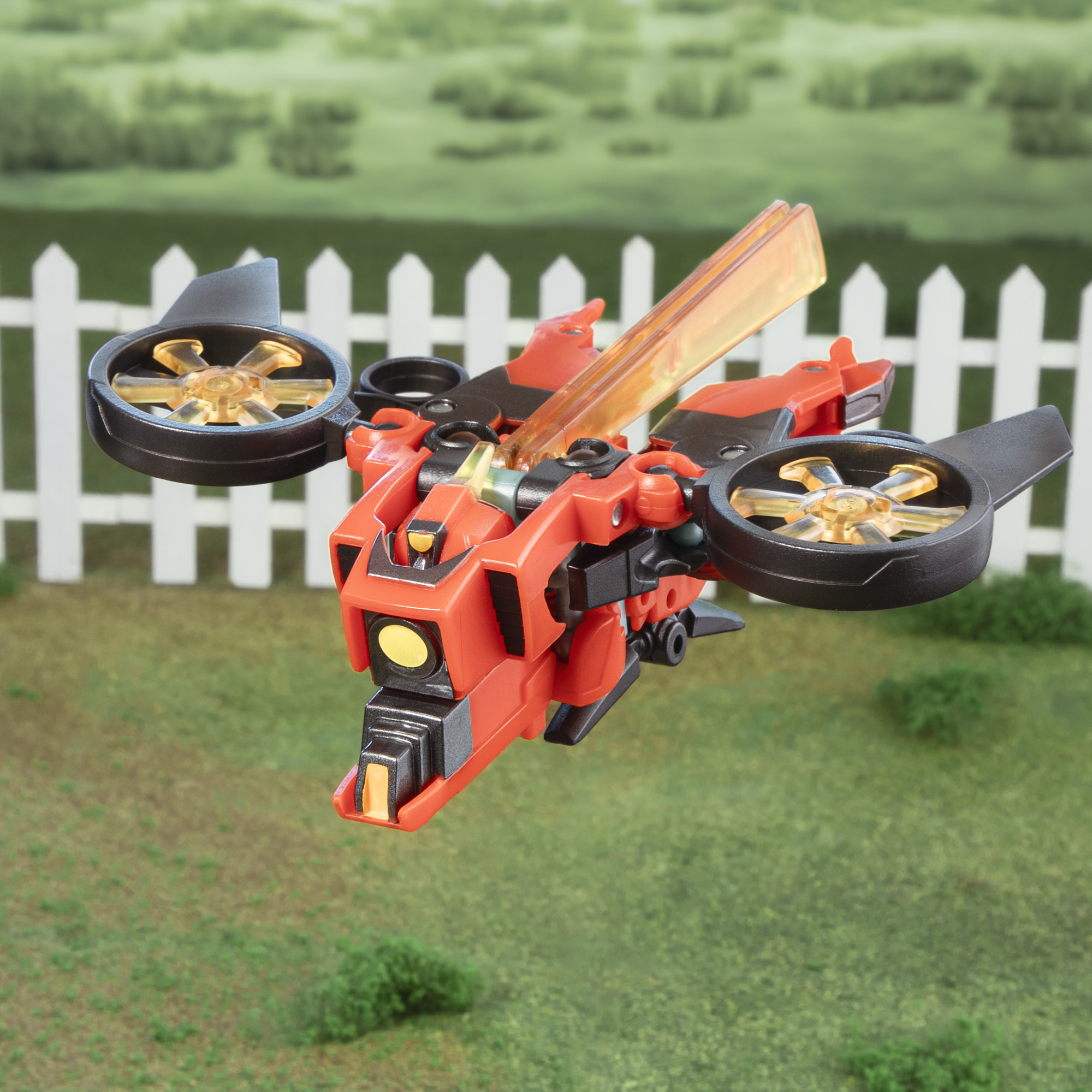 Drone giocattolo rosso e nero con due eliche, che vola sopra l'erba verde e una recinzione bianca.
