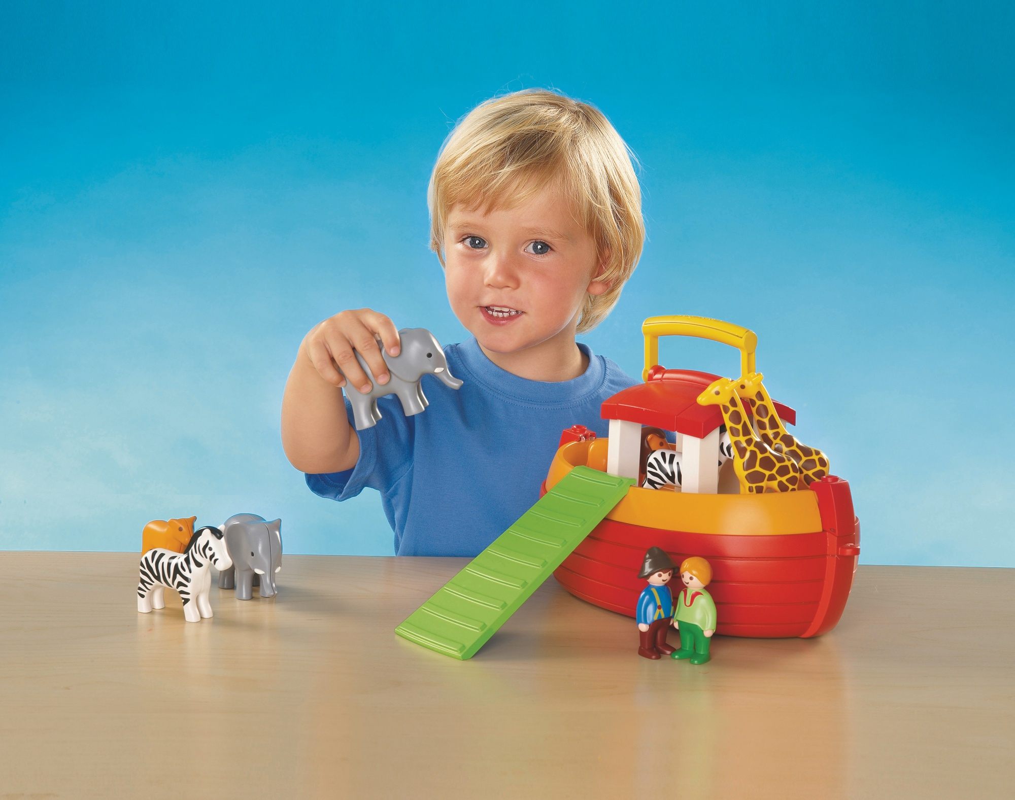Ein kleiner Junge spielt mit einer Spielzeug-Arche mit Tierfiguren auf blauem Hintergrund.