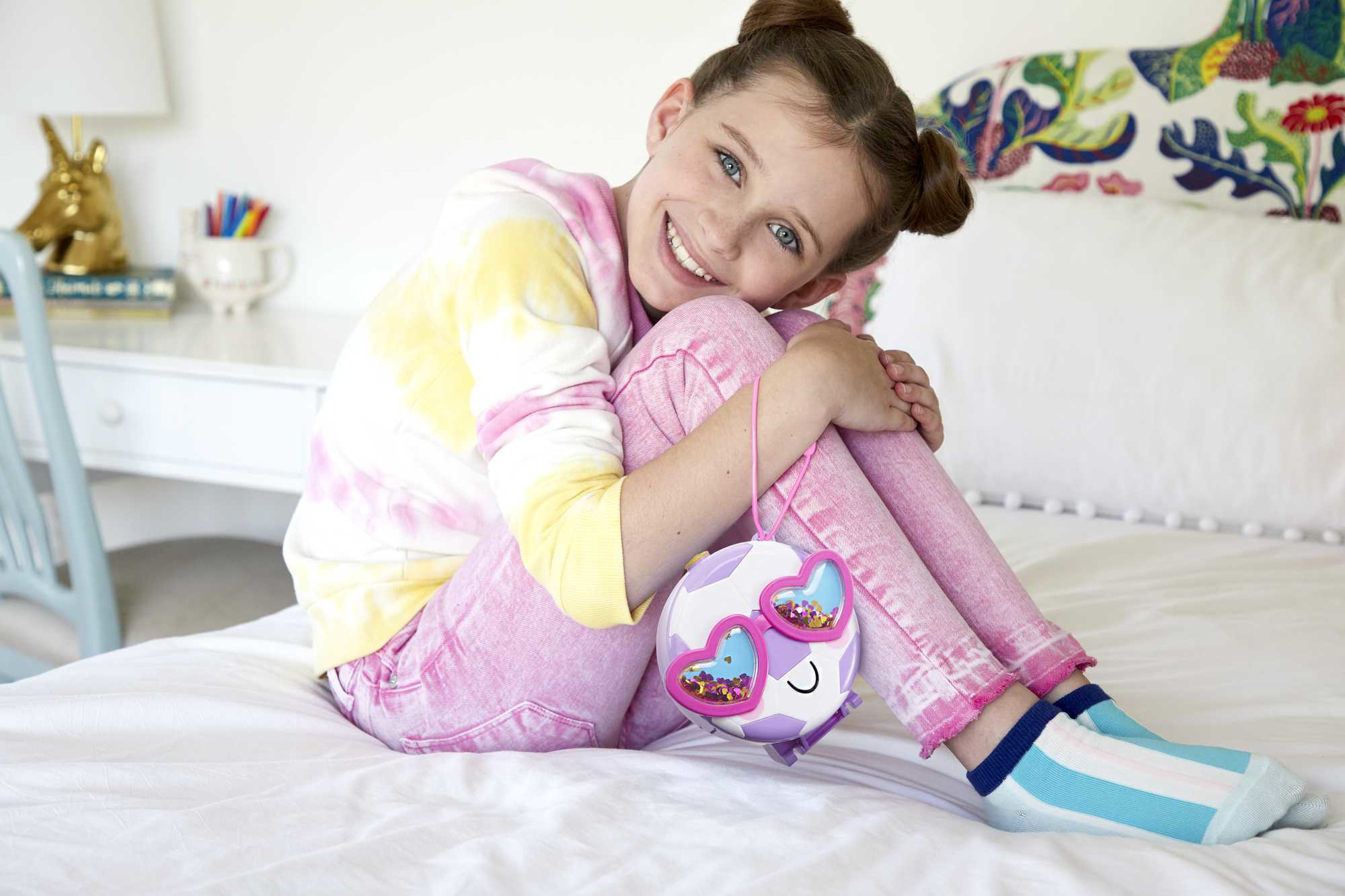 Ein lächelndes Mädchen sitzt auf einem Bett mit einer Handtasche. Sie trägt ein Batik-Shirt und pinke Hose.