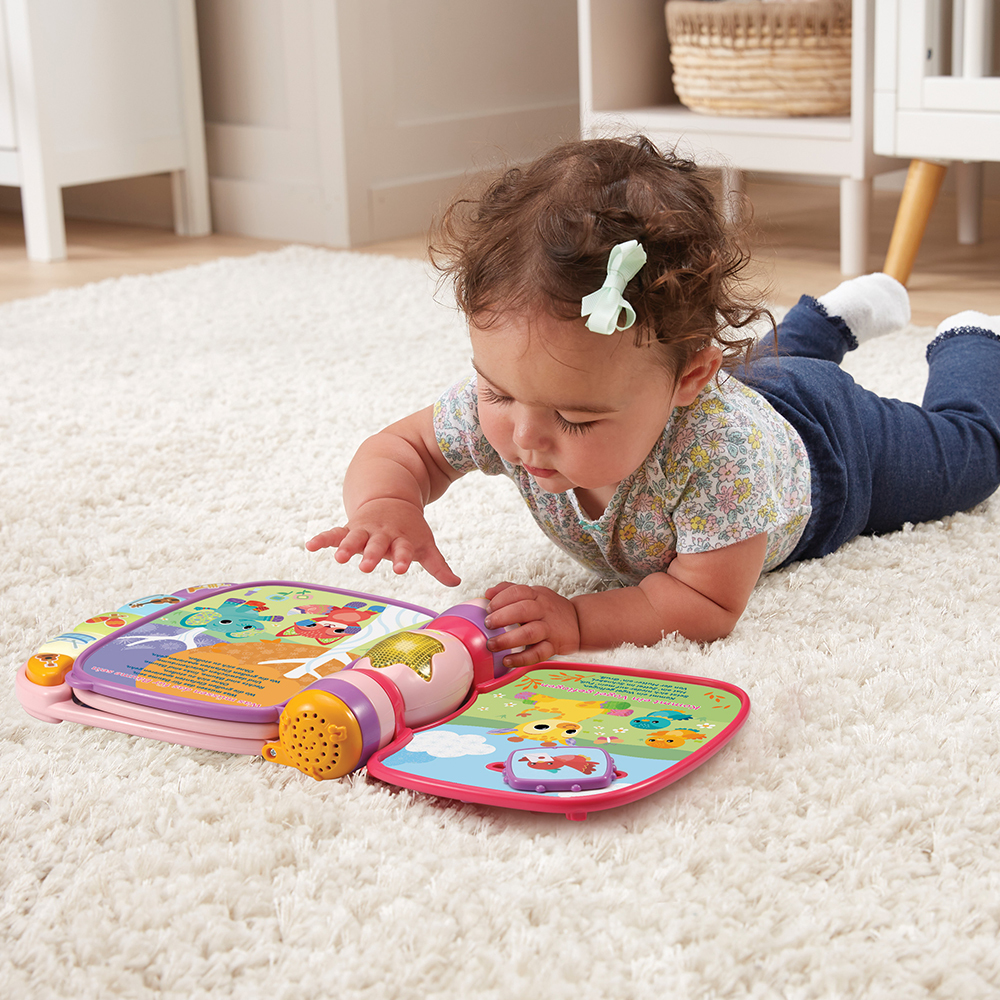 Baby auf einem Teppich, liest ein buntes Buch, greift nach einem Spielzeugmikrofon.