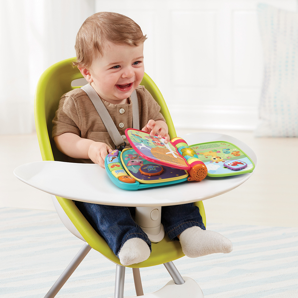 Ein Baby sitzt in einem Hochstuhl, lacht und betrachtet ein Spielzeugbuch.