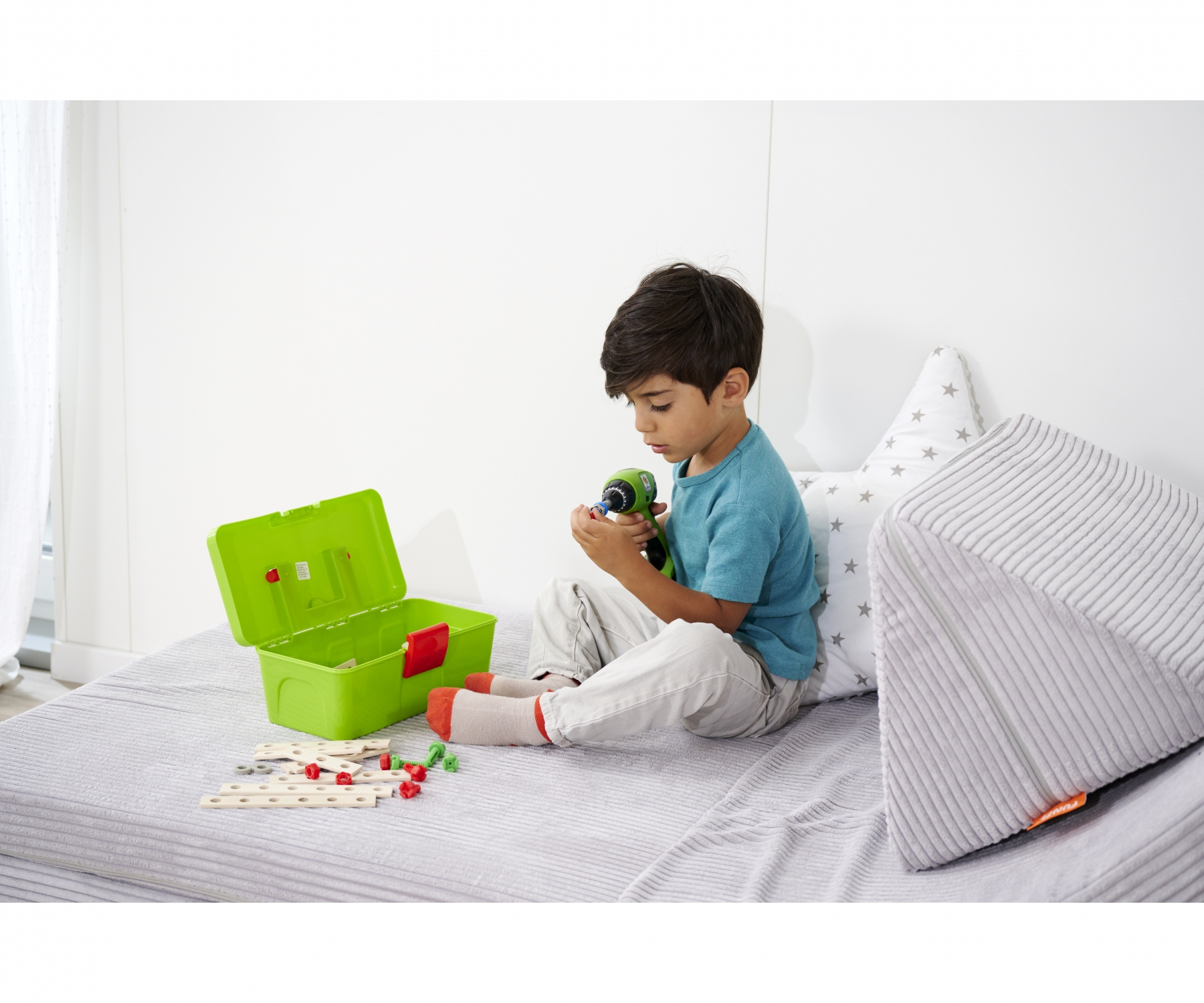 Ein kleiner Junge sitzt auf einem Bett und spielt mit einem Spielzeugbohrer. Eine grüne Werkzeugkiste und Holzstücke sind in der Nähe.