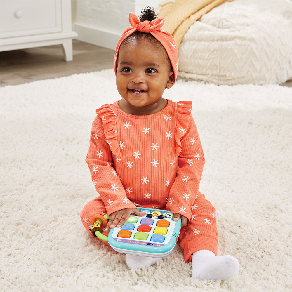 Baby sitzt auf einem weißen Teppich, hält ein Spielzeug mit bunten Knöpfen und trägt ein orangefarbenes Outfit.