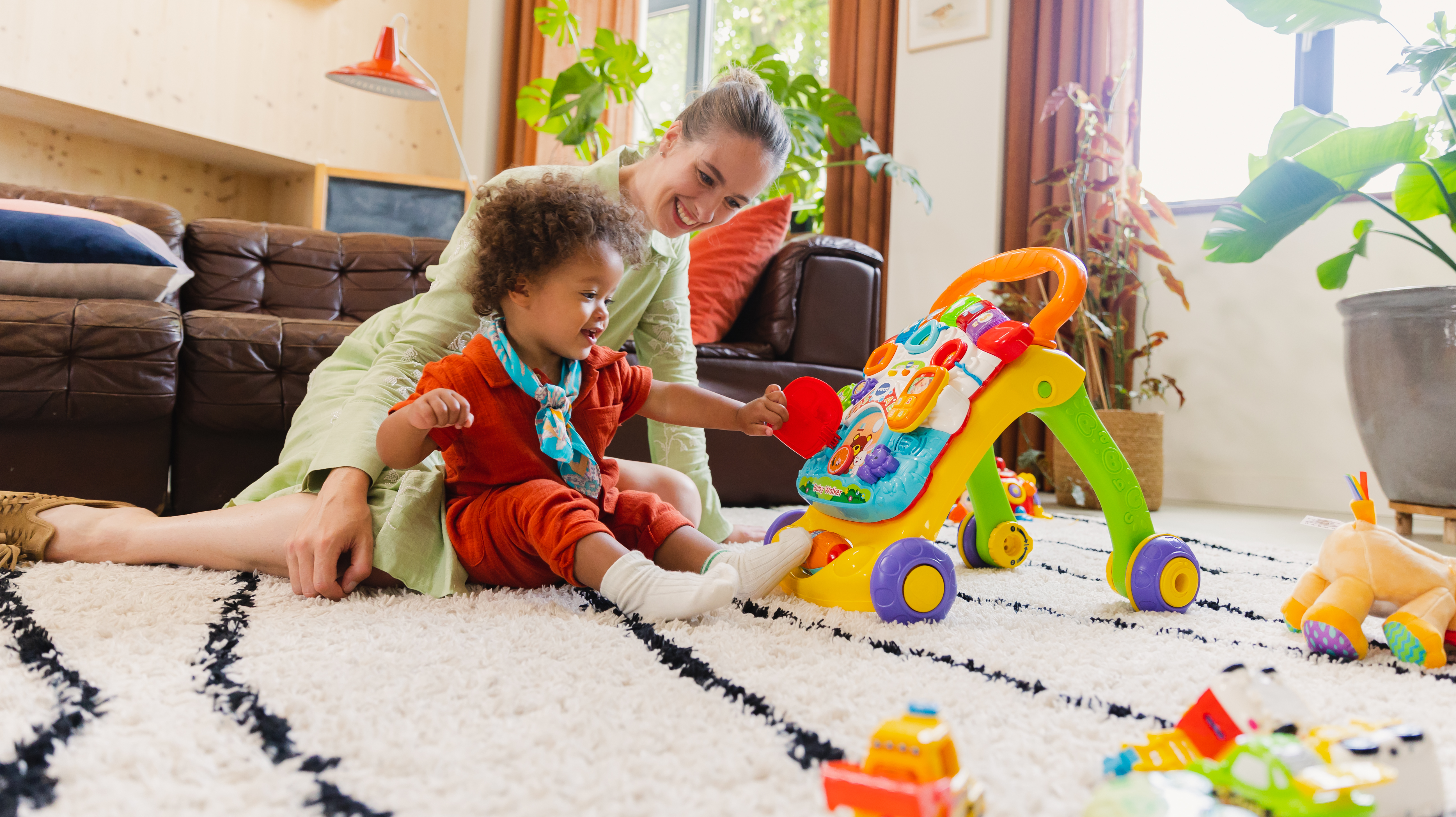 Mutter und Kind spielen auf einem Teppich mit einem Baby-Lauflernwagen und Spielzeug. Pflanzen und Möbel im Hintergrund.