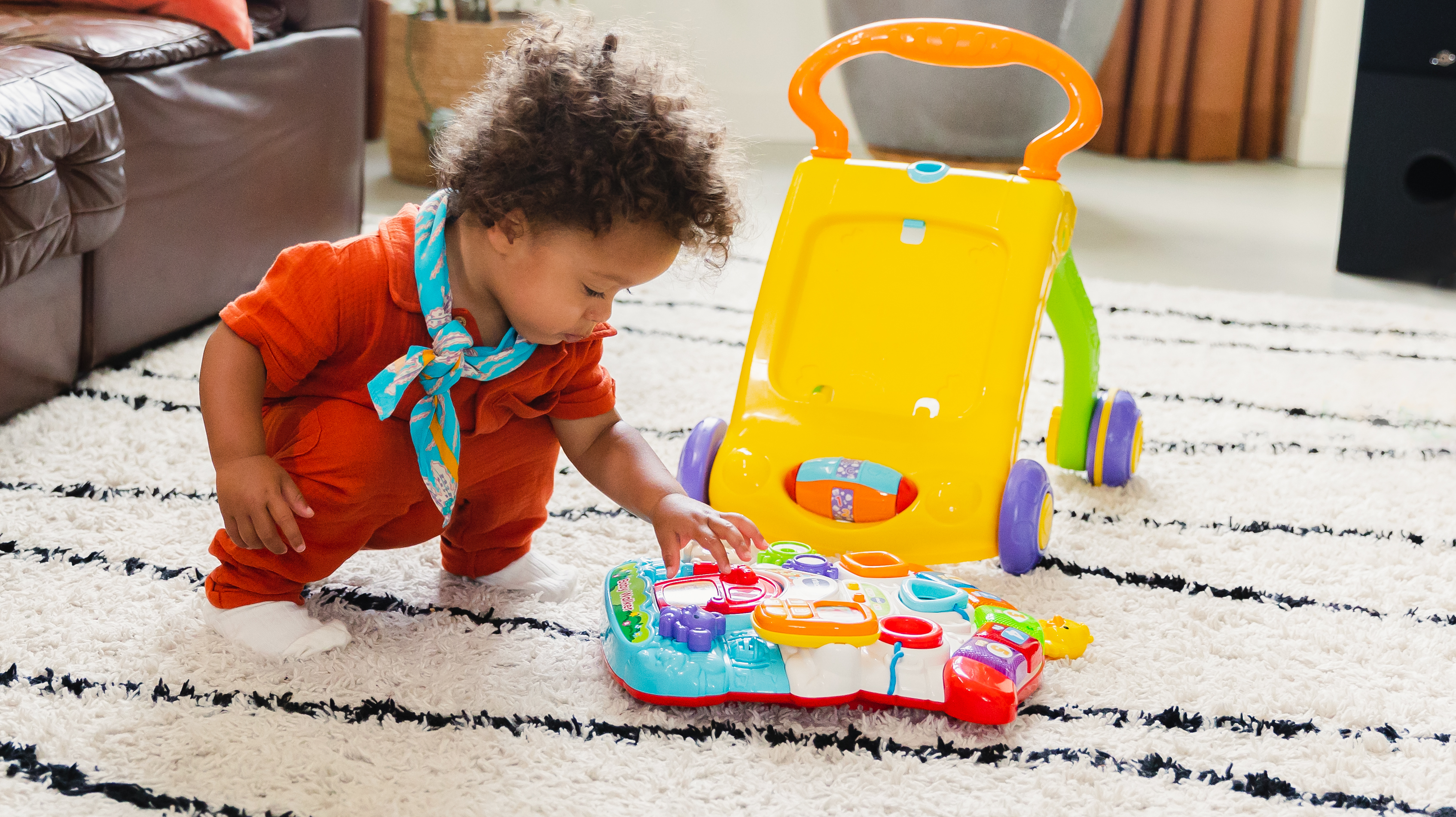 Ein Baby spielt mit einem Spielzeug auf einem Teppich. Ein gelber Lauflerner steht hinter dem Baby.