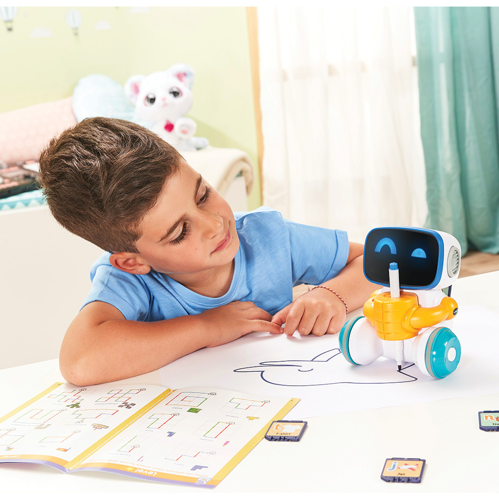 Ein Junge betrachtet einen Roboter auf einem Tisch. Der Roboter hat einen Bildschirm und ist gelb und blau. Eine Spielzeugkatze ist im Hintergrund.