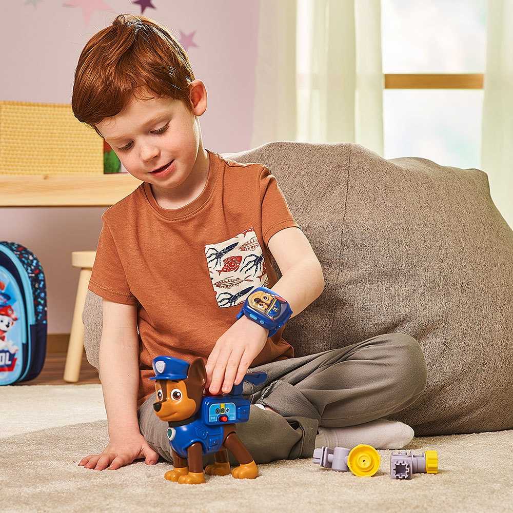 Ein Junge spielt mit einem Spielzeughund, der eine Uhr trägt. Paw Patrol Rucksack und Spielzeuge sind im Bild.