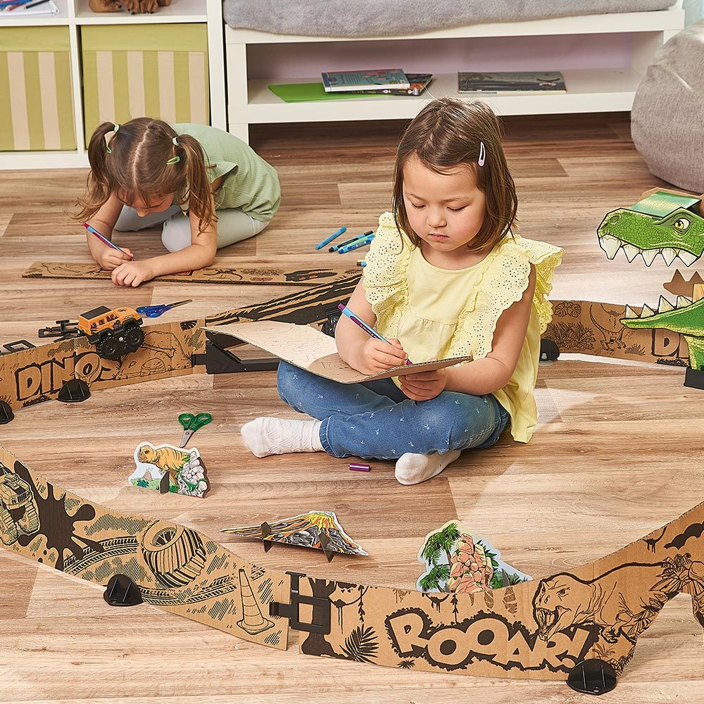 Zwei Mädchen zeichnen auf Papp-Dinosaurier-Streckenabschnitten, umgeben von Spielzeug-Dinosauriern. Sie sind auf einem Holzboden.