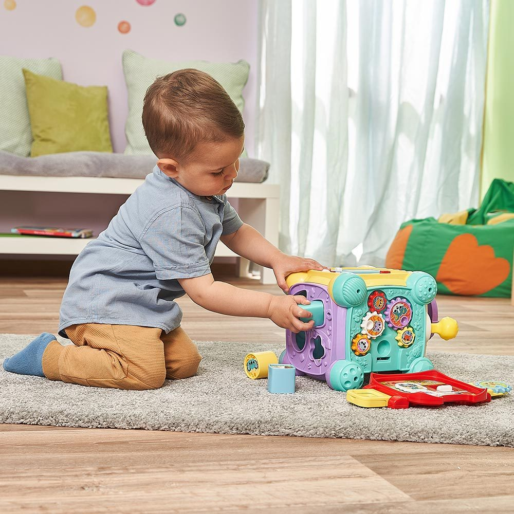 Ein Baby spielt mit einem Würfelspielzeug mit verschiedenen Formen und Zahnrädern auf einem Teppich.