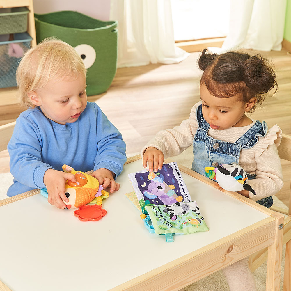 Zwei Kleinkinder spielen mit Spielzeug. Eines hält ein Stoffbuch, das andere ein Stofftier.