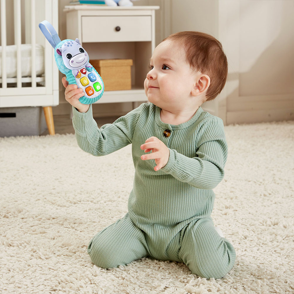 Baby hält Spielzeugtelefon, trägt grünen Strampler, auf Teppich.