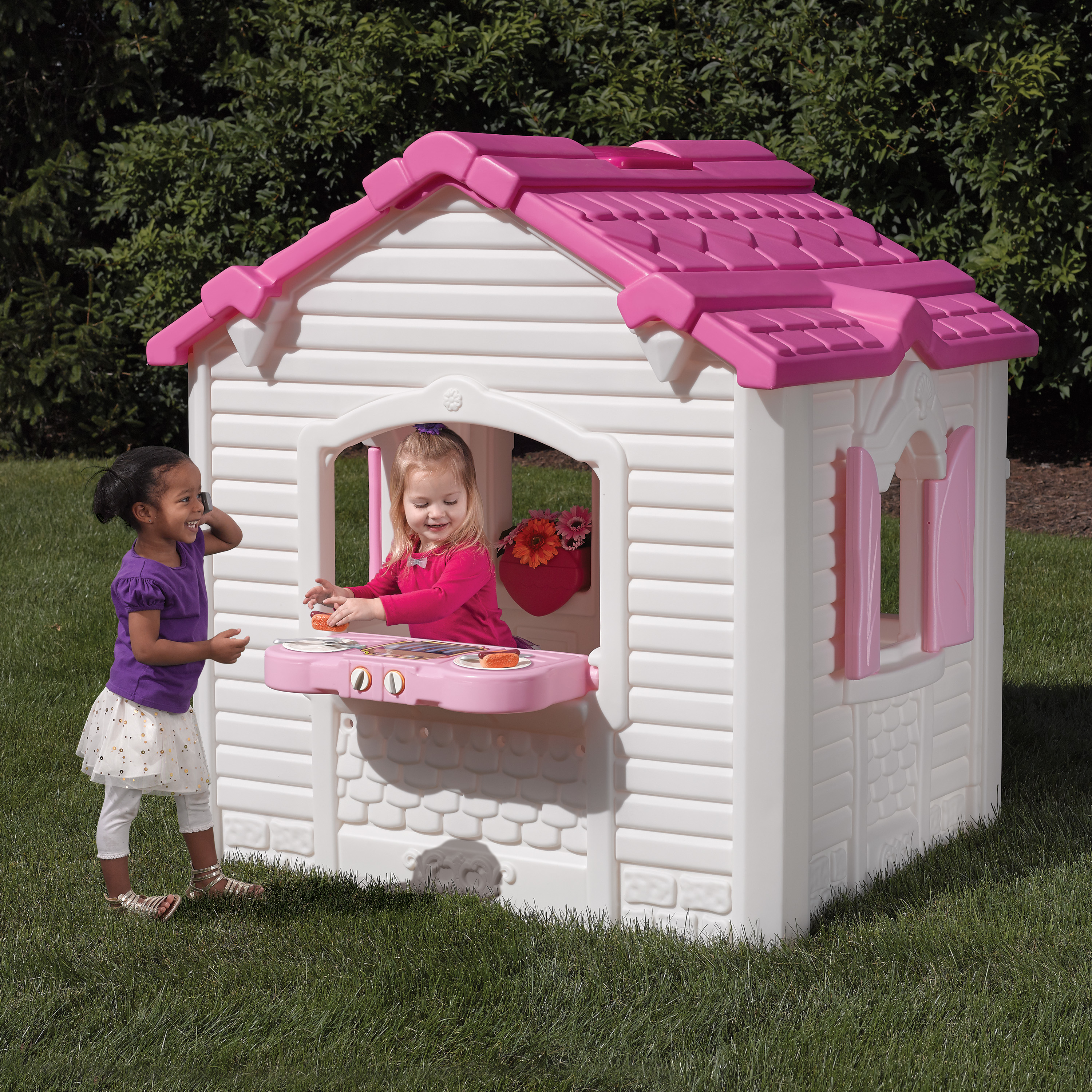 Zwei Mädchen spielen in einem rosa-weißen Spielhaus auf Gras. Blumen im Fenster.