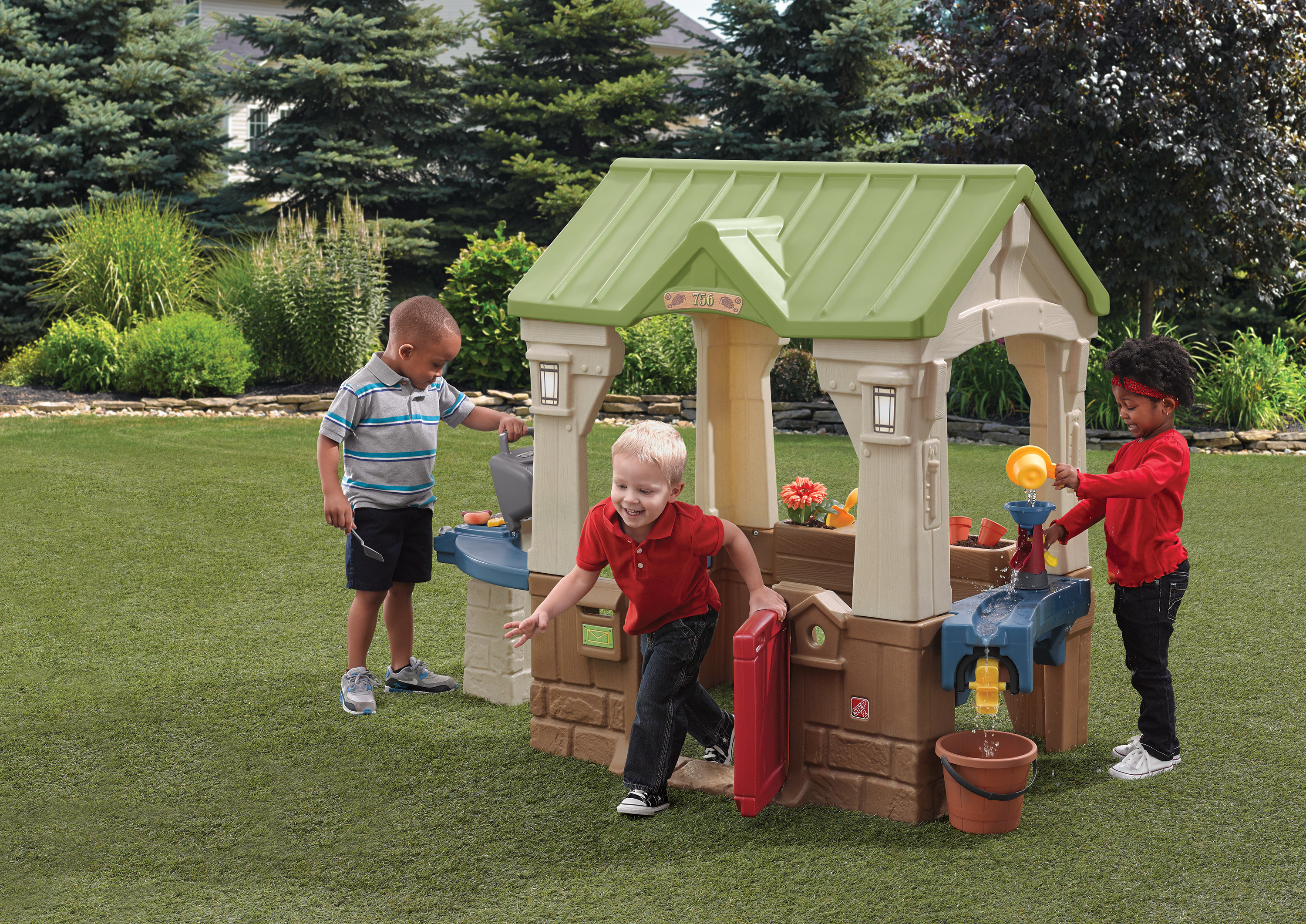 Drei Kinder spielen in einem Spielhaus. Das Spielhaus ist beige und grün, auf grünem Gras.