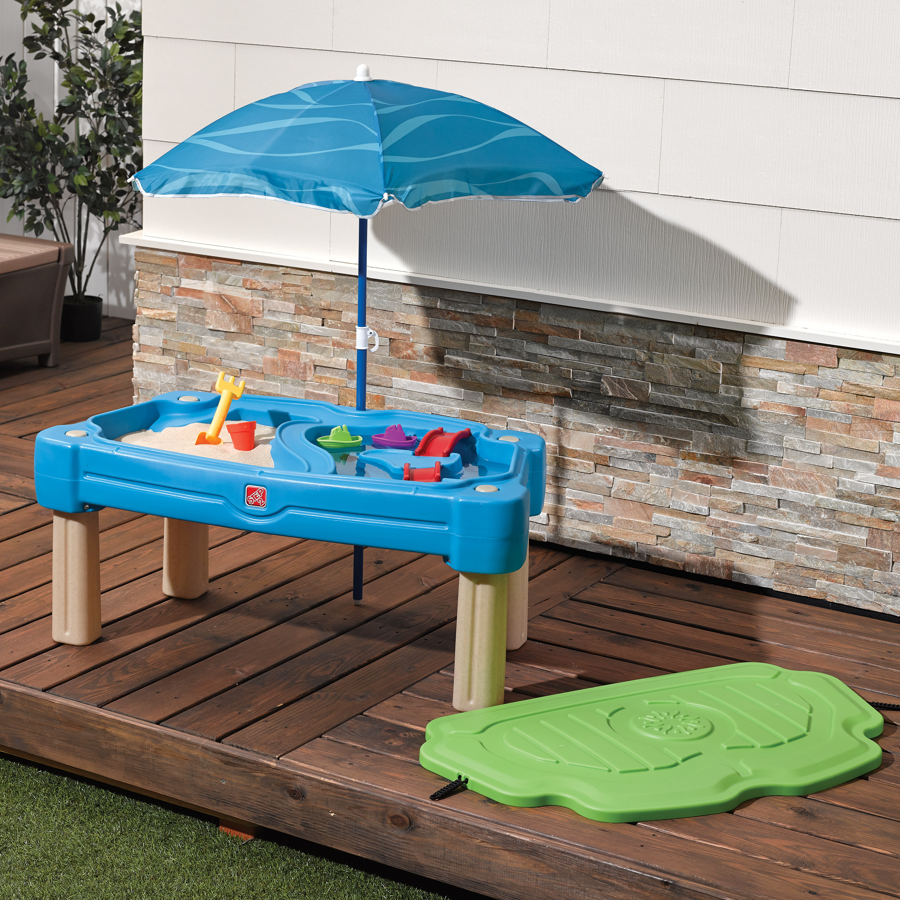 Blauer Sandkasten-Tisch mit Schirm, auf Holzterrasse, daneben grüner Deckel.
