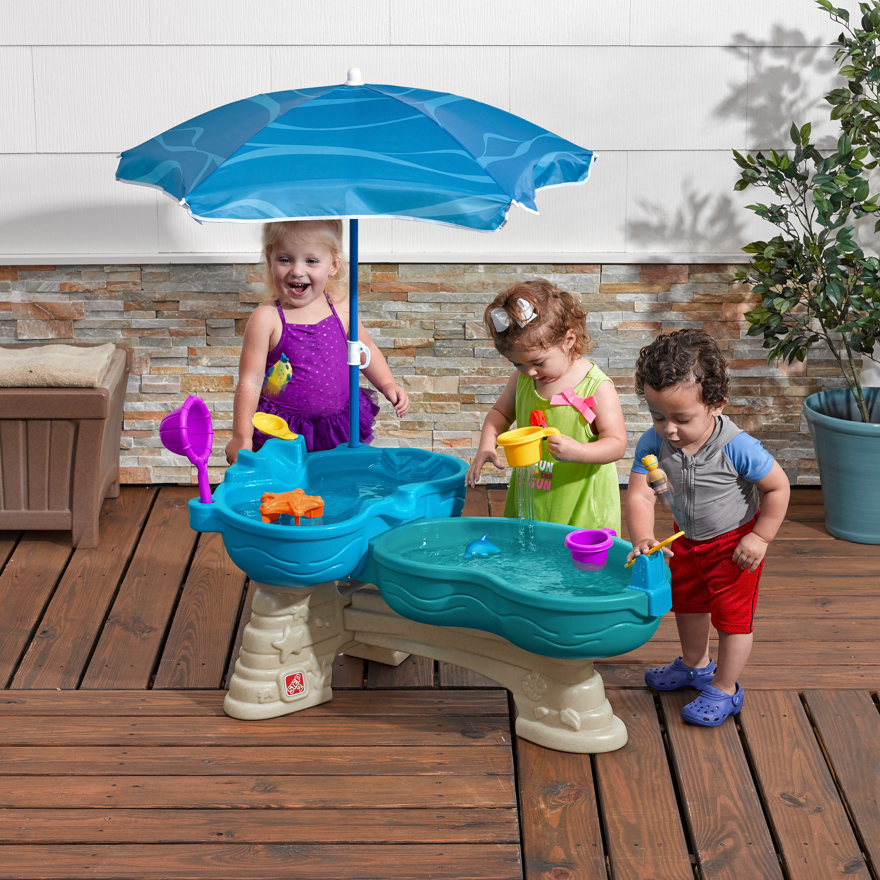 Drei Kinder spielen mit Wasser in einem blau-beige Wassertisch unter einem blauen Sonnenschirm.