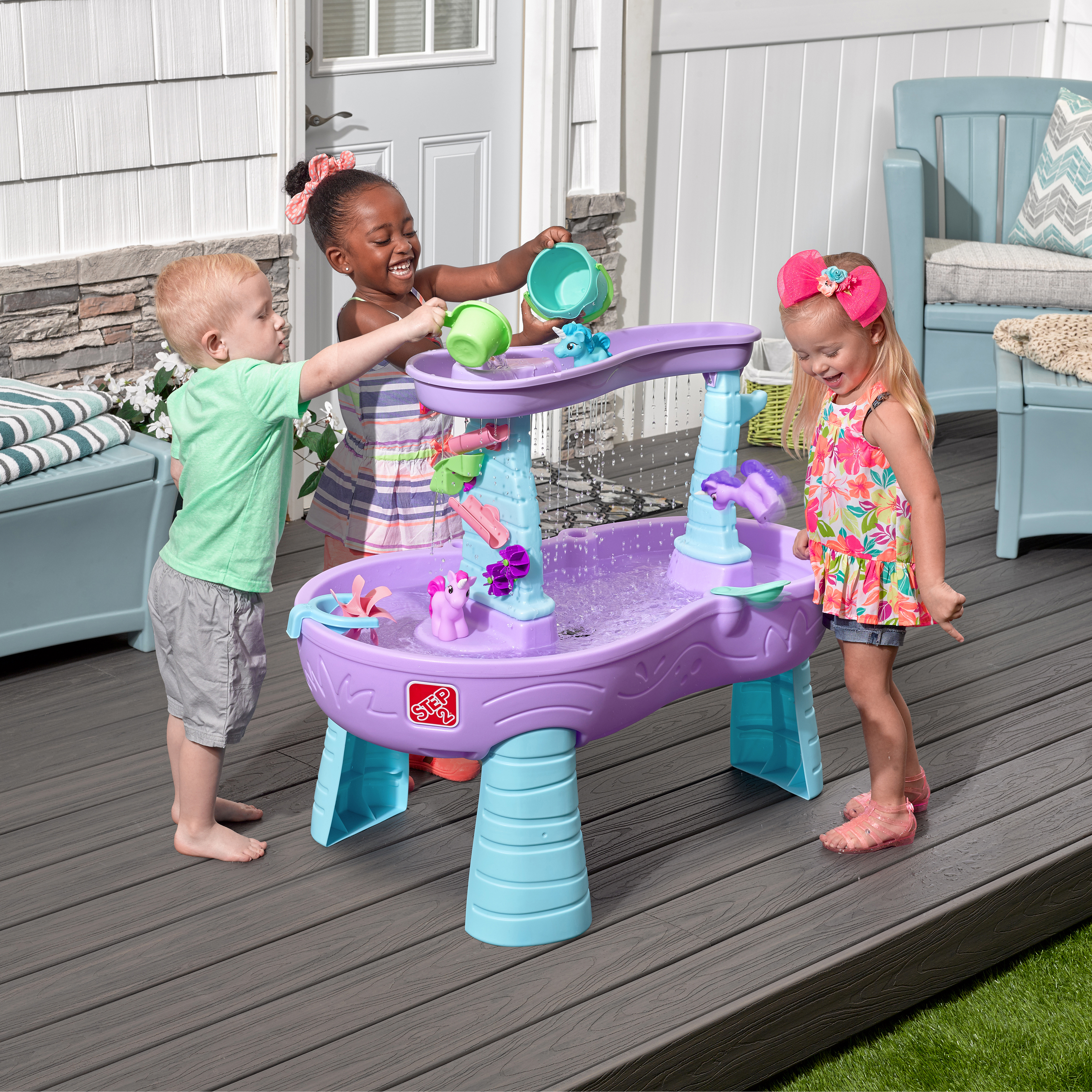 Drei Kinder spielen mit einem lila Wassertisch auf einem Deck und gießen Wasser.