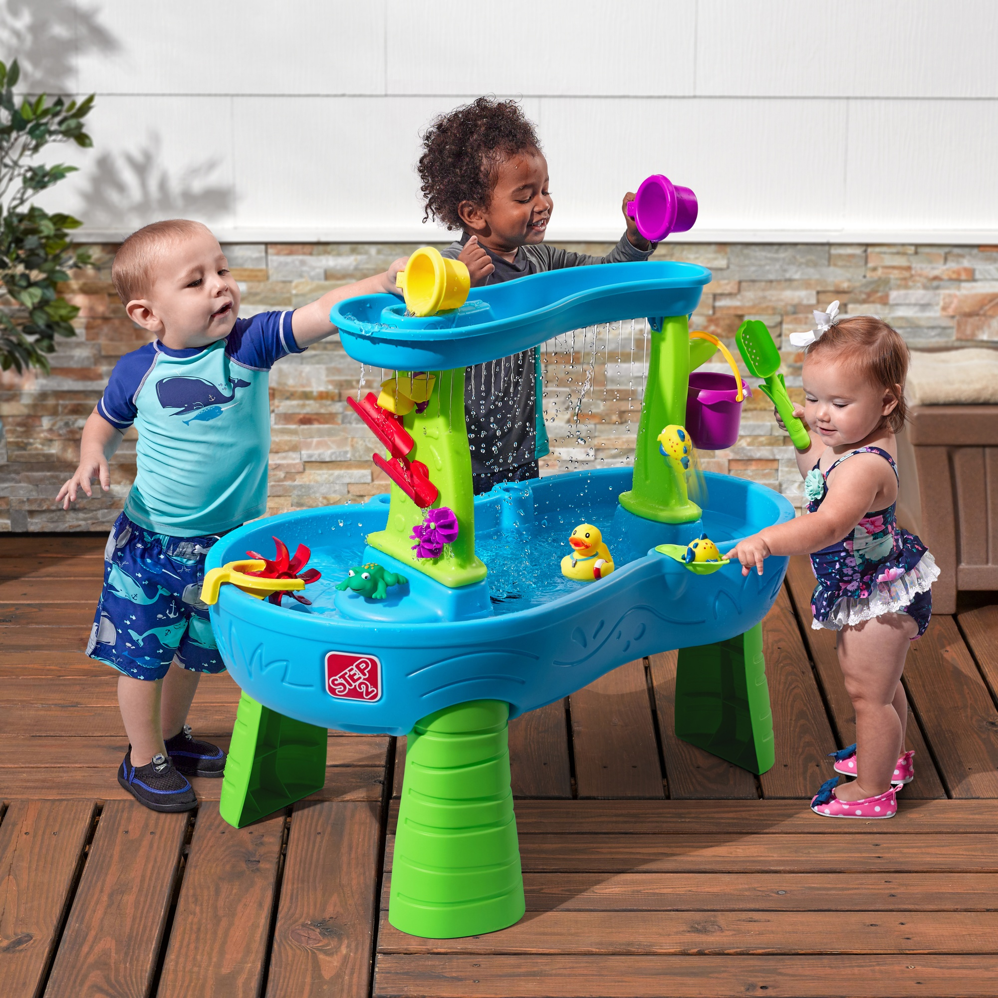Drei Kinder spielen draußen mit einem blau-grünen Wassertisch und Spielzeug.