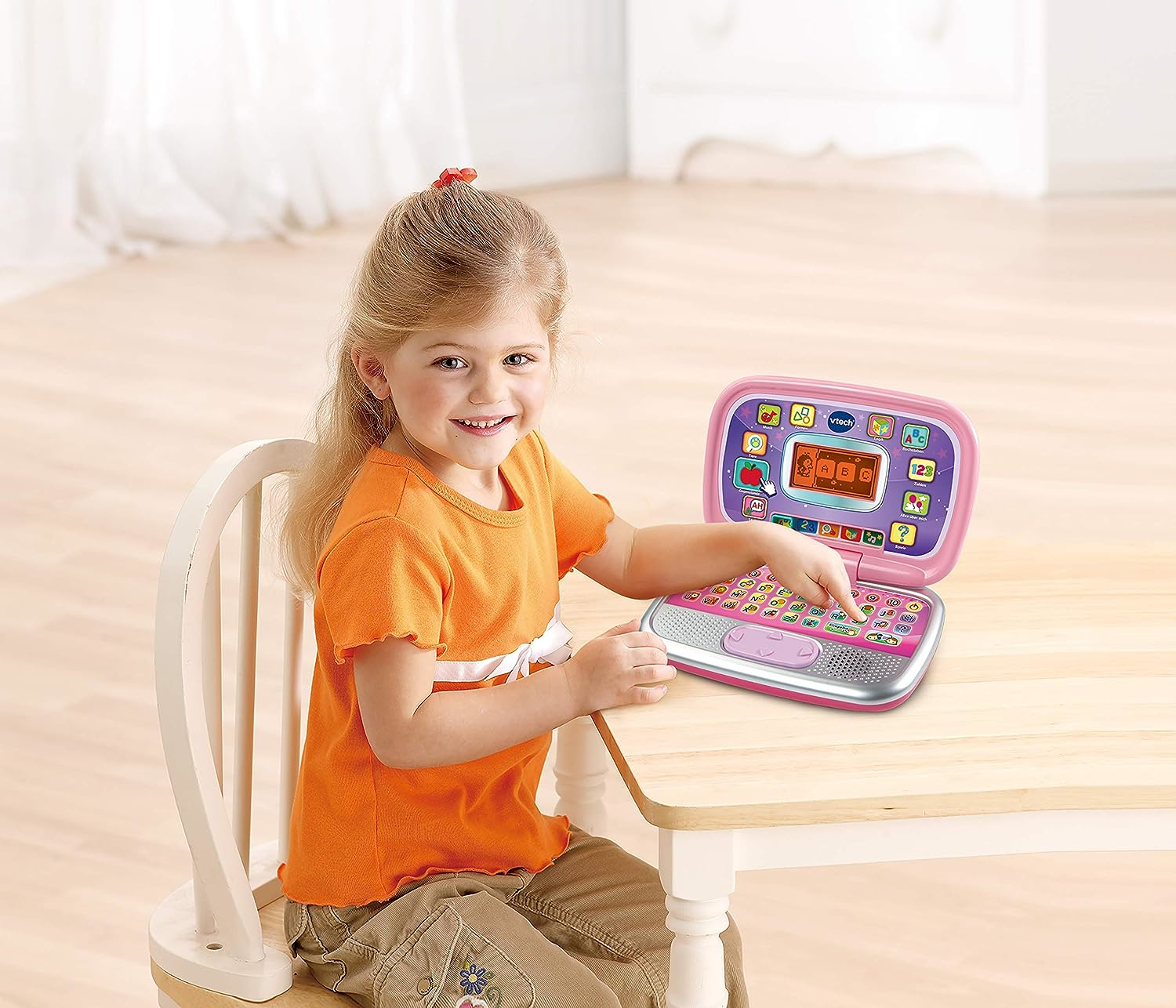 Ein Mädchen sitzt an einem Tisch und benutzt einen rosa Spielzeug-Laptop, lächelt, mit einem Stuhl links.