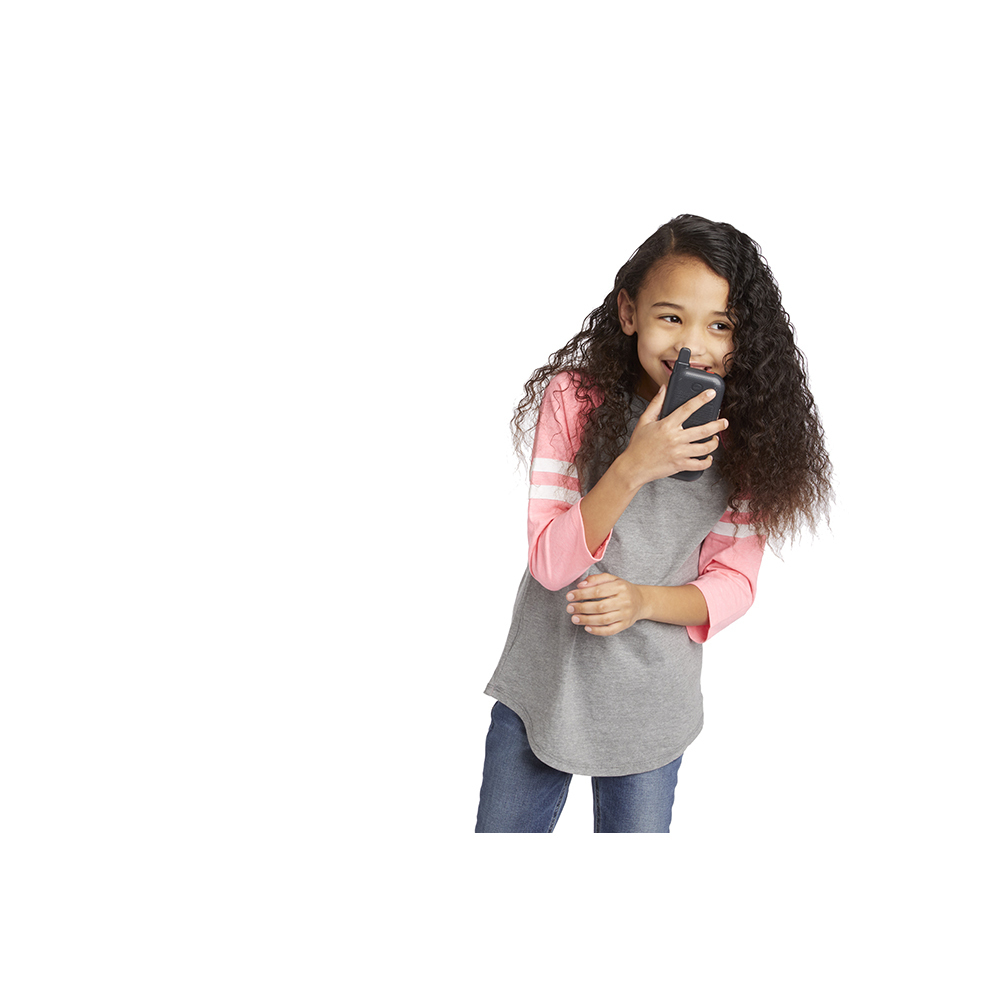 Ein Mädchen mit lockigem Haar hält ein schwarzes Walkie-Talkie und lächelt. Sie trägt ein graues und rosa Hemd und blaue Jeans.