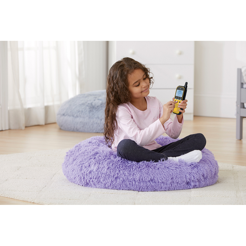Ein Mädchen sitzt auf einem lila, pelzigen Kissen und hält ein Walkie-Talkie, lächelnd. Eine Kommode im Hintergrund.