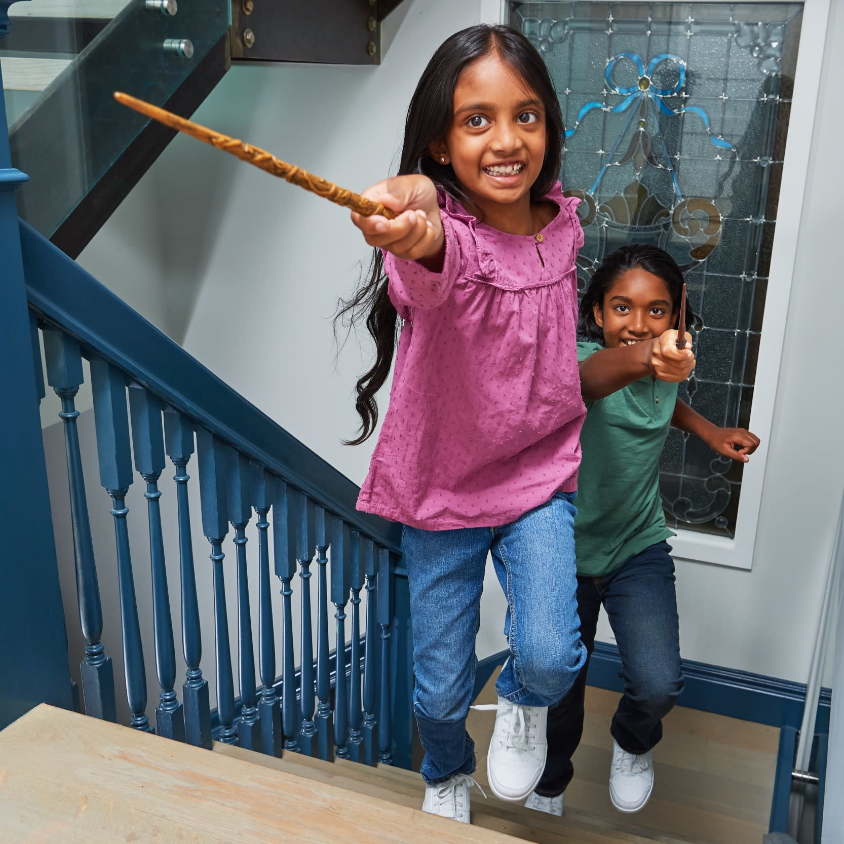 Dwie dziewczyny biegną po schodach trzymając różdżki. Dziewczyna z przodu się uśmiecha. Schody mają niebieskie boki.