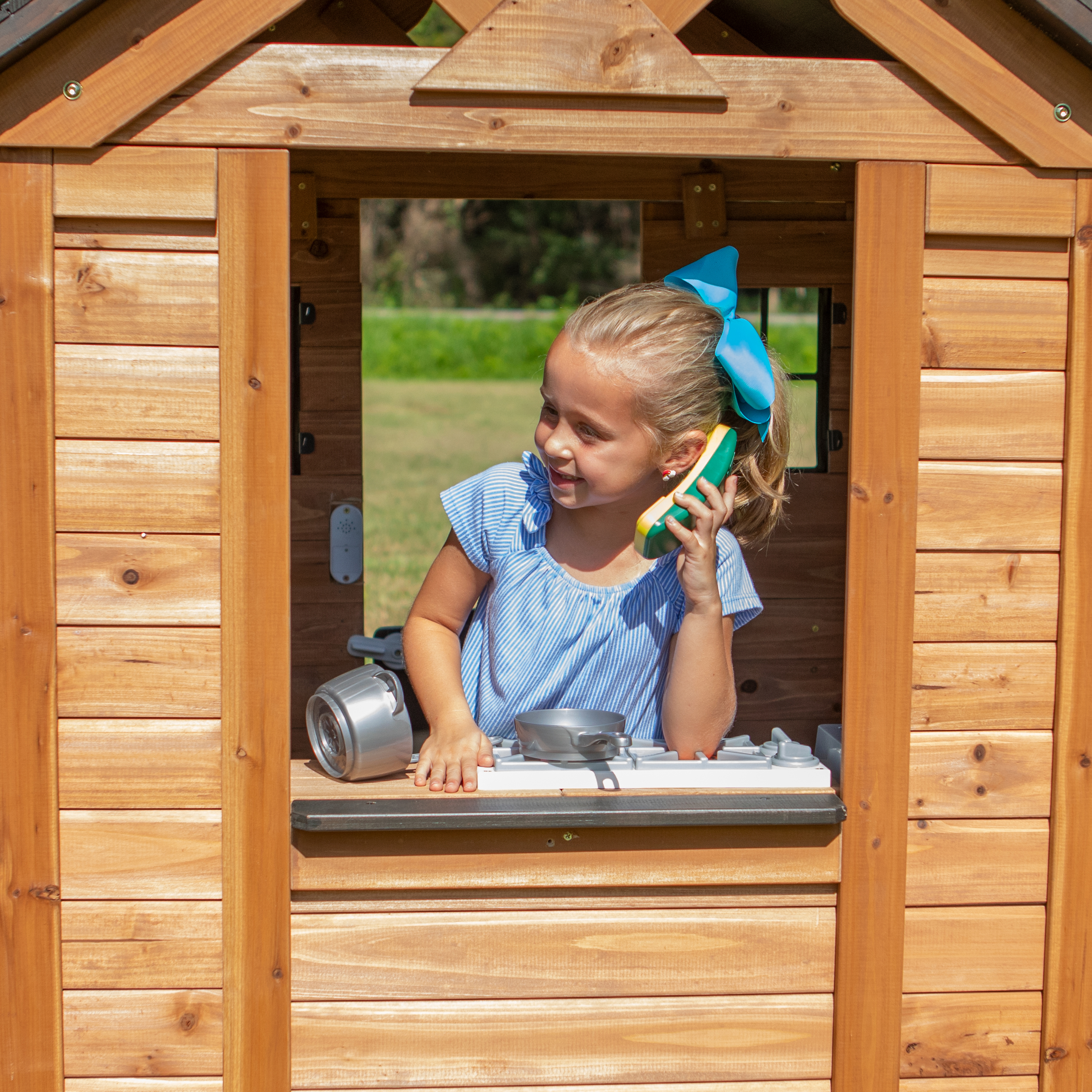 Ein Mädchen in einem Holzspielhaus-Fenster, das mit einem Spielzeugtelefon spricht, mit einer Küche im Inneren.