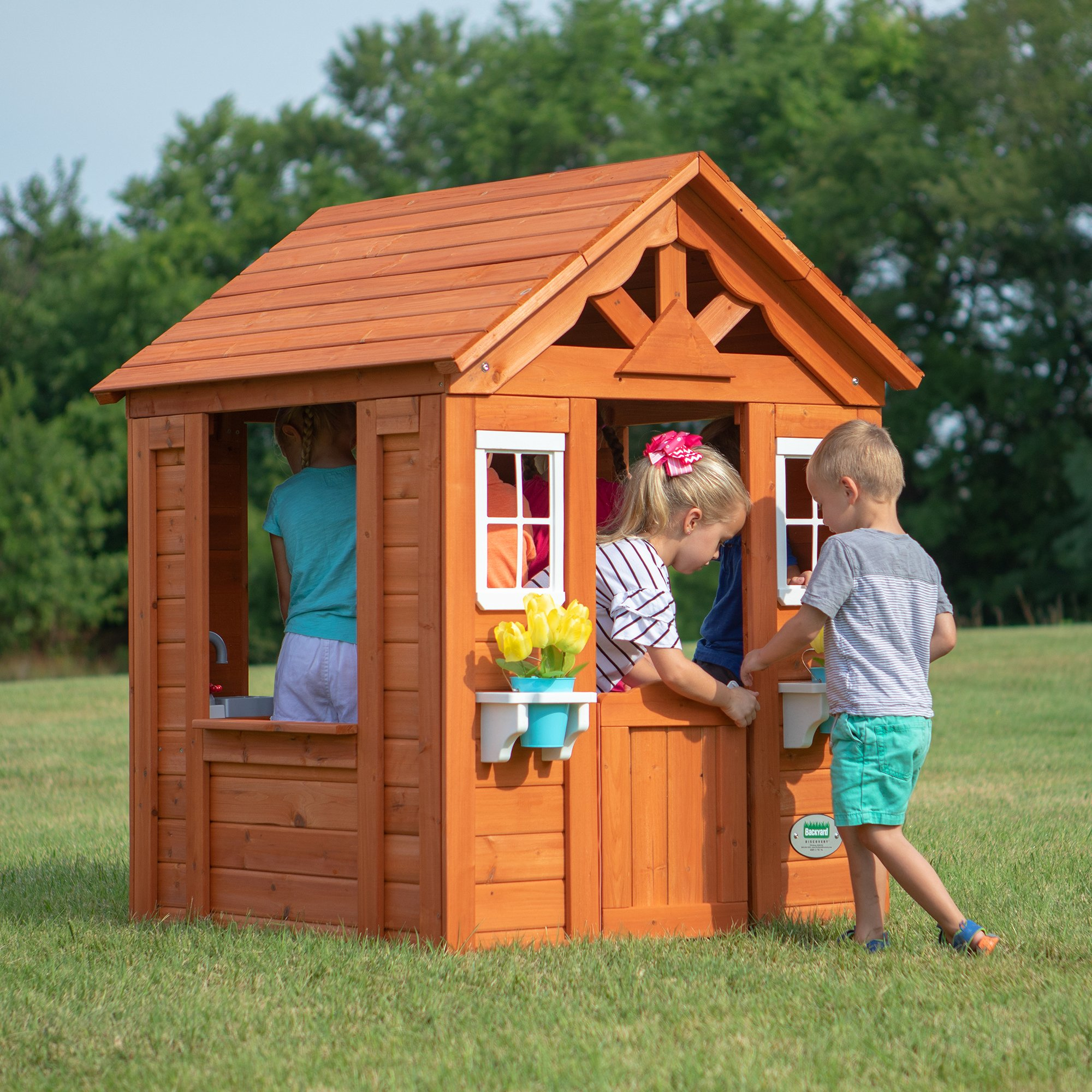 Ein hölzernes Spielhaus auf einer Wiese. Kinder spielen in der Nähe der Tür und Fenster.