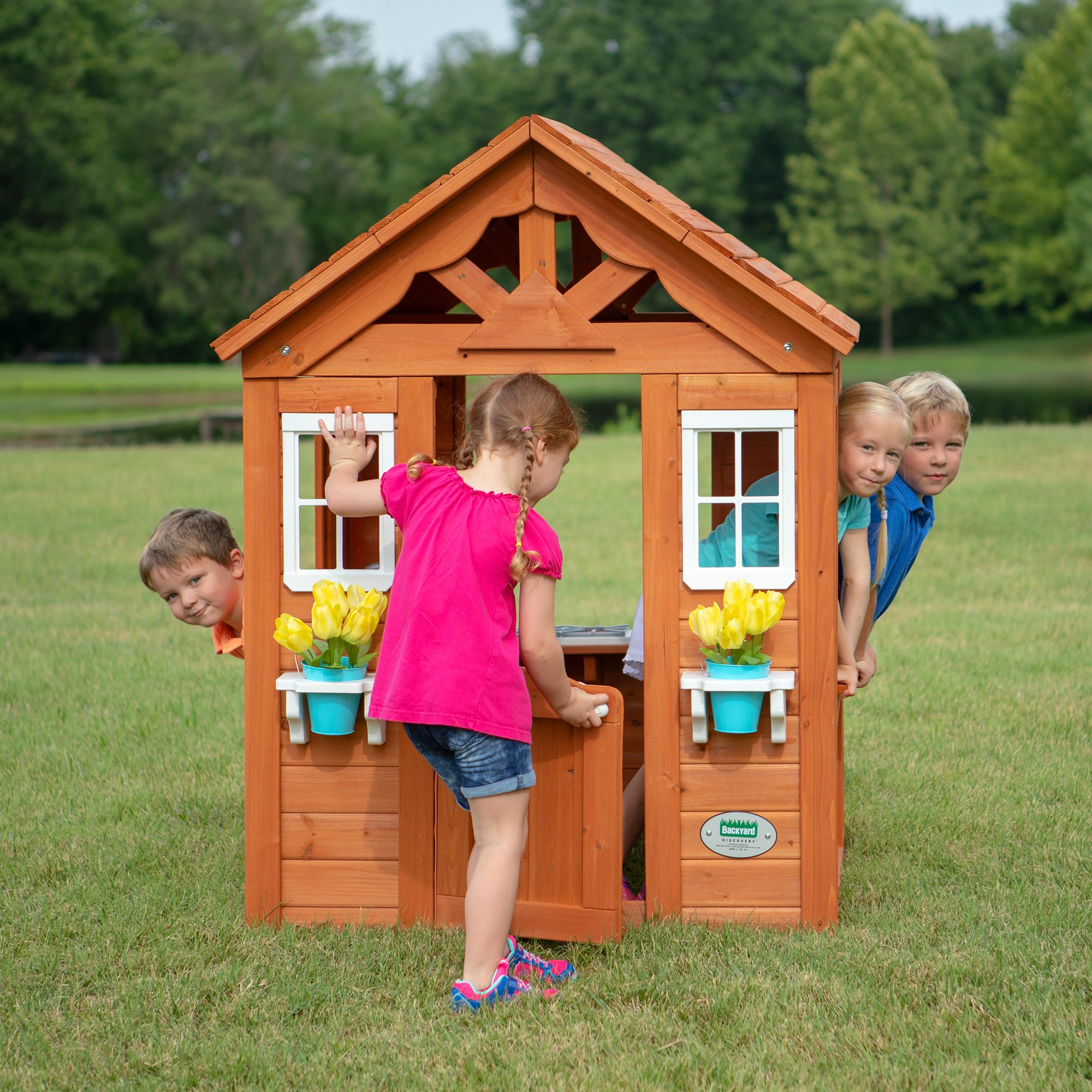 Drei Kinder spielen in einem Holzspielhaus mit Blumentöpfen. Sie sind draußen auf dem Rasen.