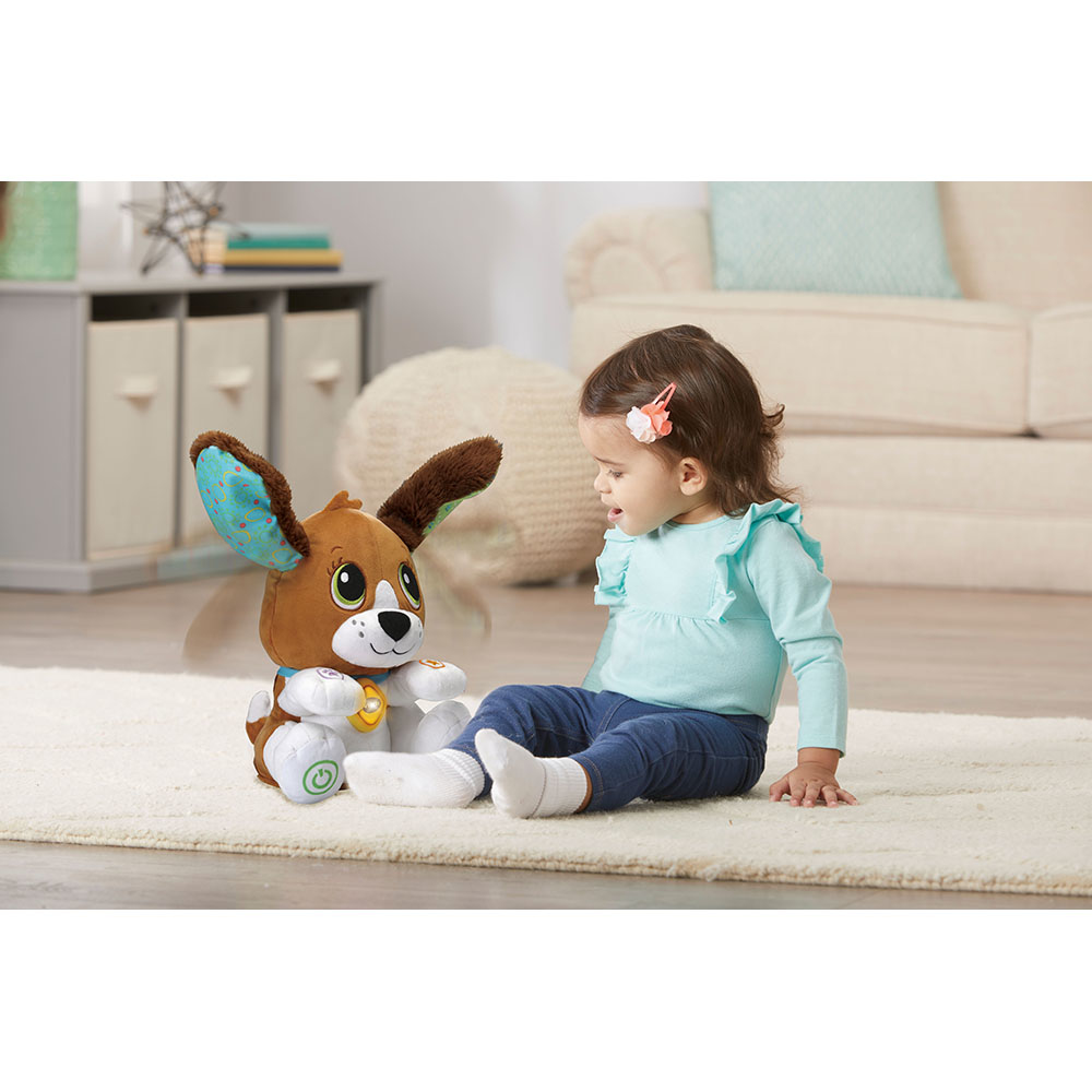 Ein Baby sitzt mit einem Spielzeug-Welpen auf einem Teppich und schaut es an.
