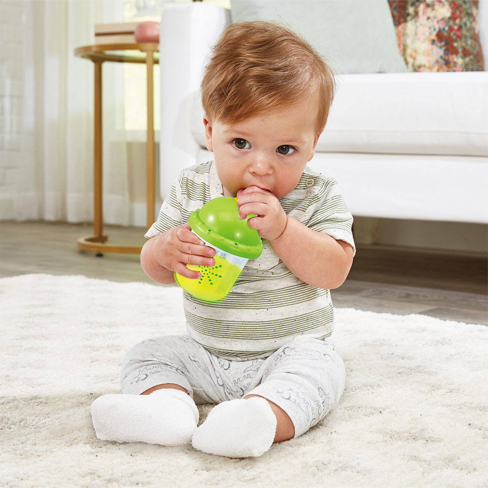 Baby hält grünen Trinkbecher, sitzt auf weißem Teppich, vor einem Sofa.