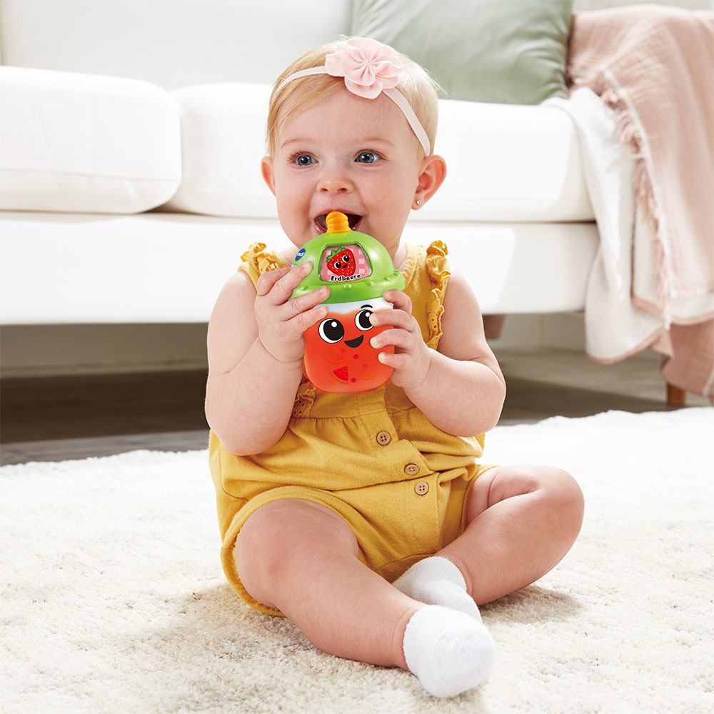 Baby mit Spielzeug, trägt Stirnband, sitzt auf einem Teppich.
