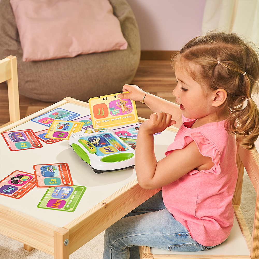 Ein junges Mädchen sitzt an einem Tisch und spielt mit Lernkarten. Verschiedene farbenfrohe Karten und ein Gerät sind sichtbar.
