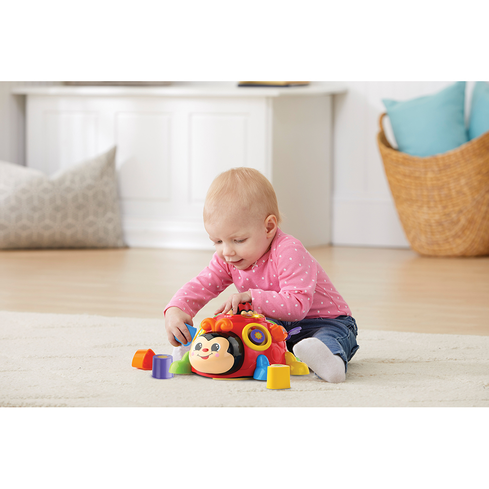 Ein Baby sitzt auf einem Teppich und spielt mit einem Marienkäfer-Spielzeug und bunten Blöcken.