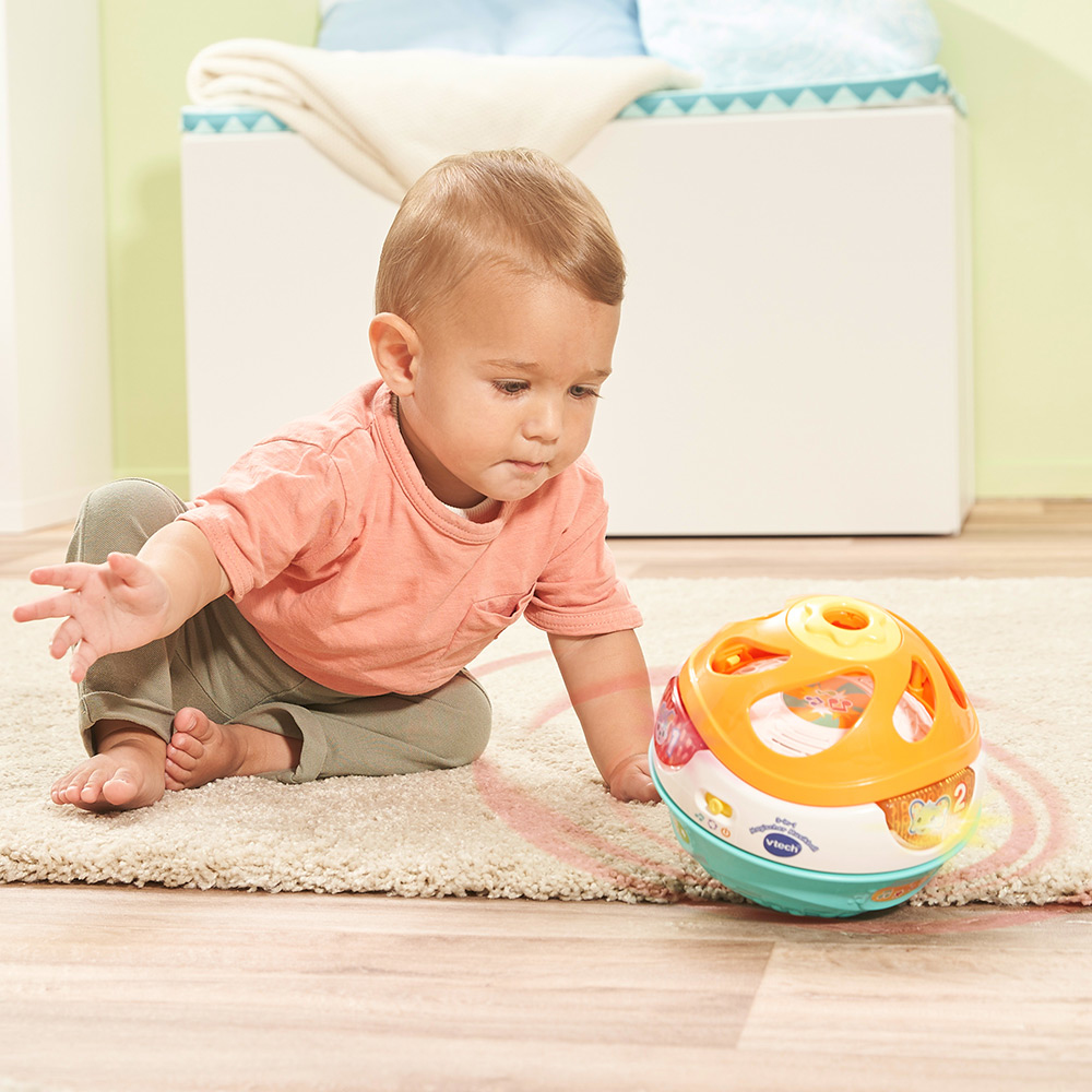 Ein Baby greift nach einem bunten, interaktiven Spielzeugball auf einem hellen Teppich.