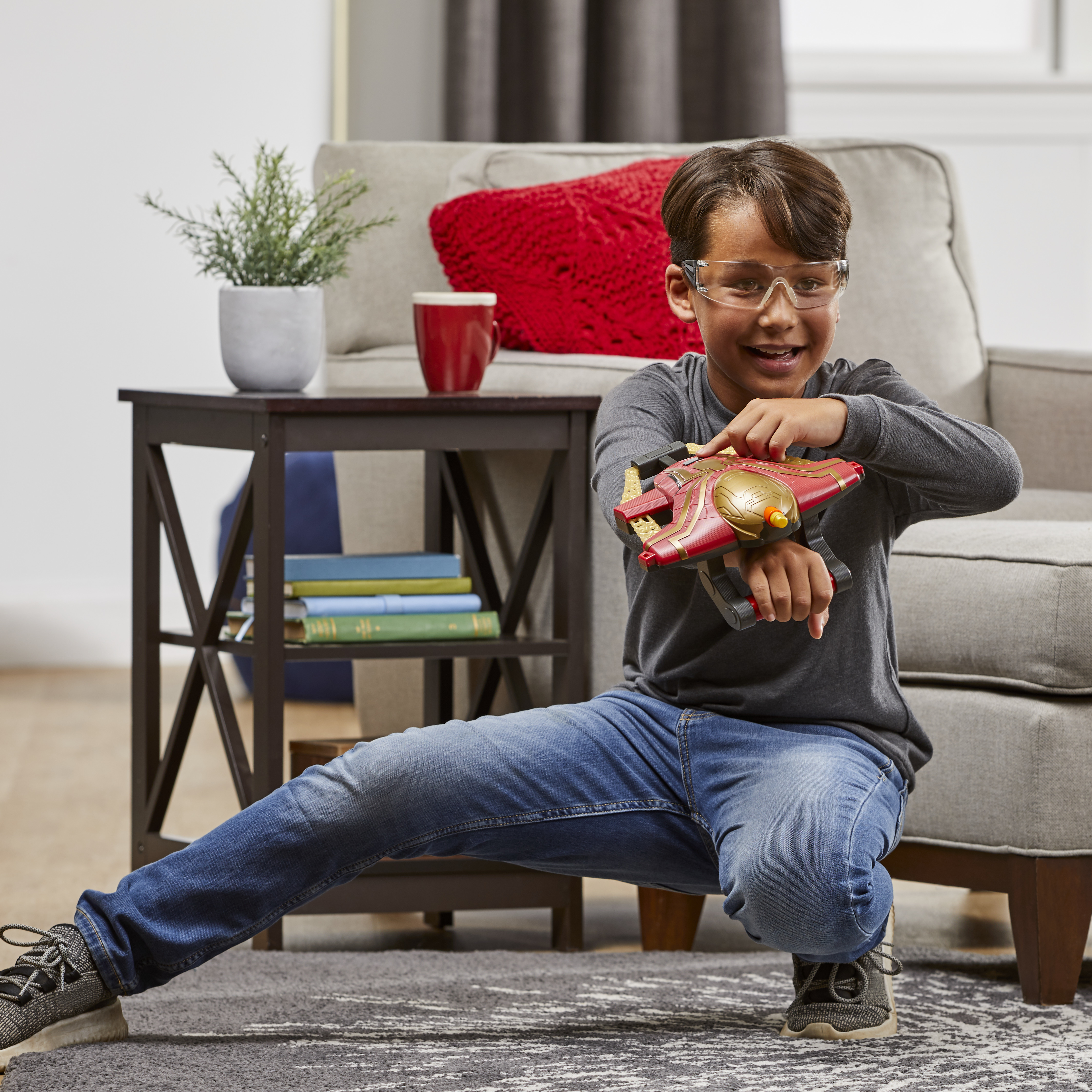 Chłopiec bawi się pistoletem zabawką. Ma na sobie gogle i kuca obok kanapy.