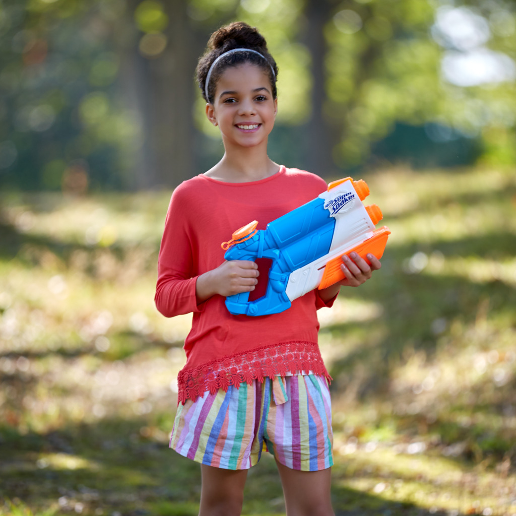 Una niña sonriente sosteniendo una pistola de agua azul y naranja en un entorno al aire libre.