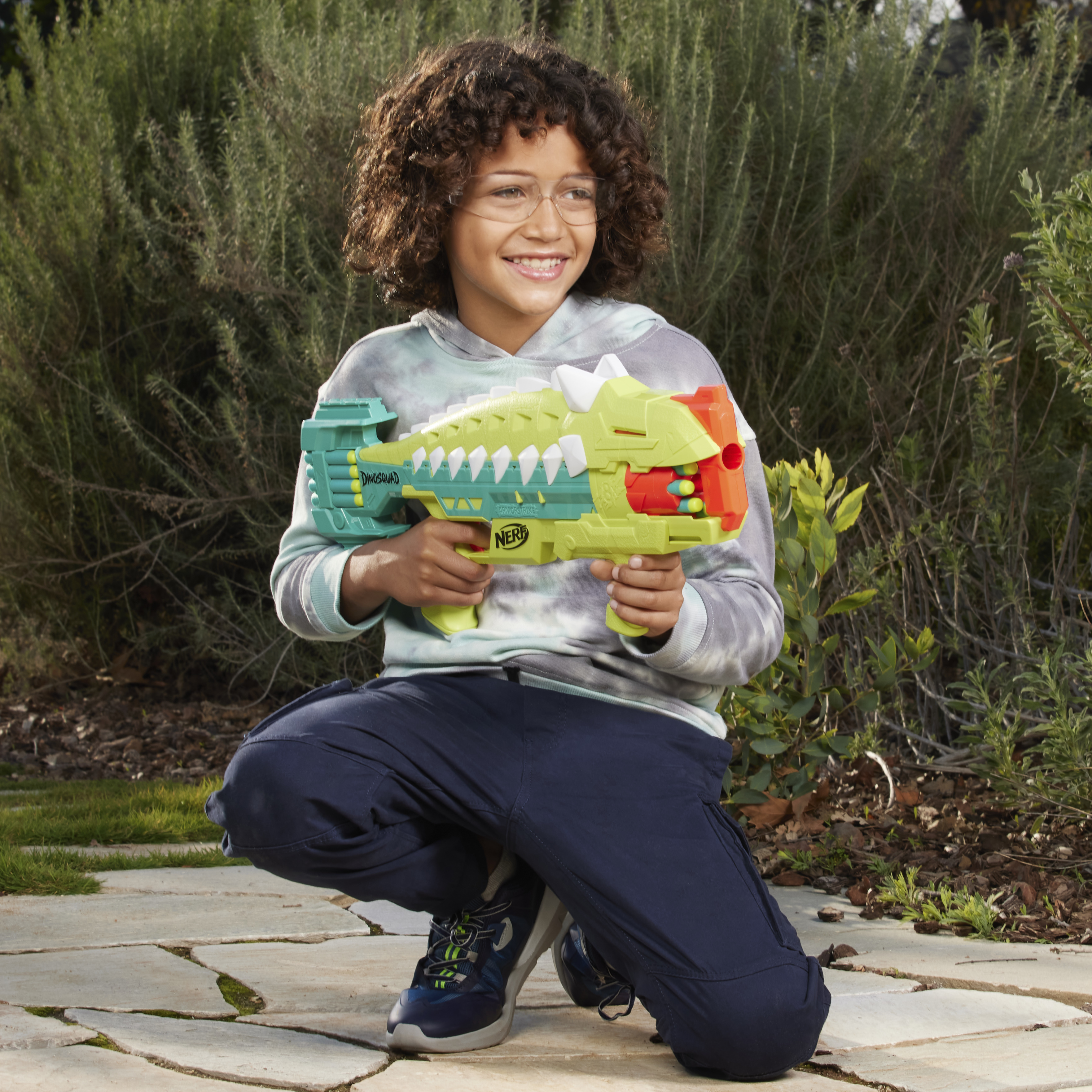 Un niño con gafas, arrodillado y sosteniendo una pistola de juguete Nerf en un jardín.