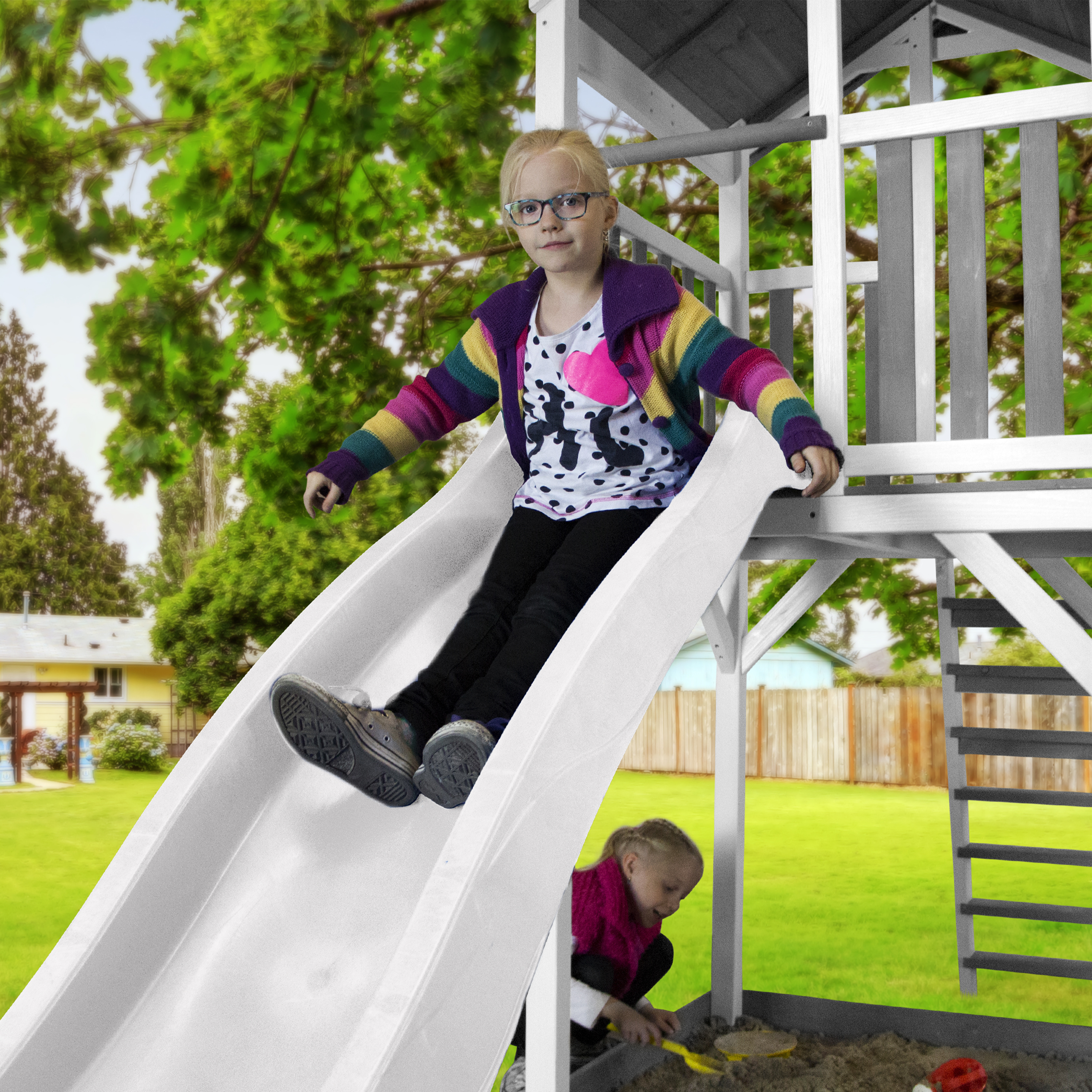 Zwei Mädchen rutschen eine weiße Rutsche auf einem Spielplatz hinunter, mit einem grünen Garten im Hintergrund.
