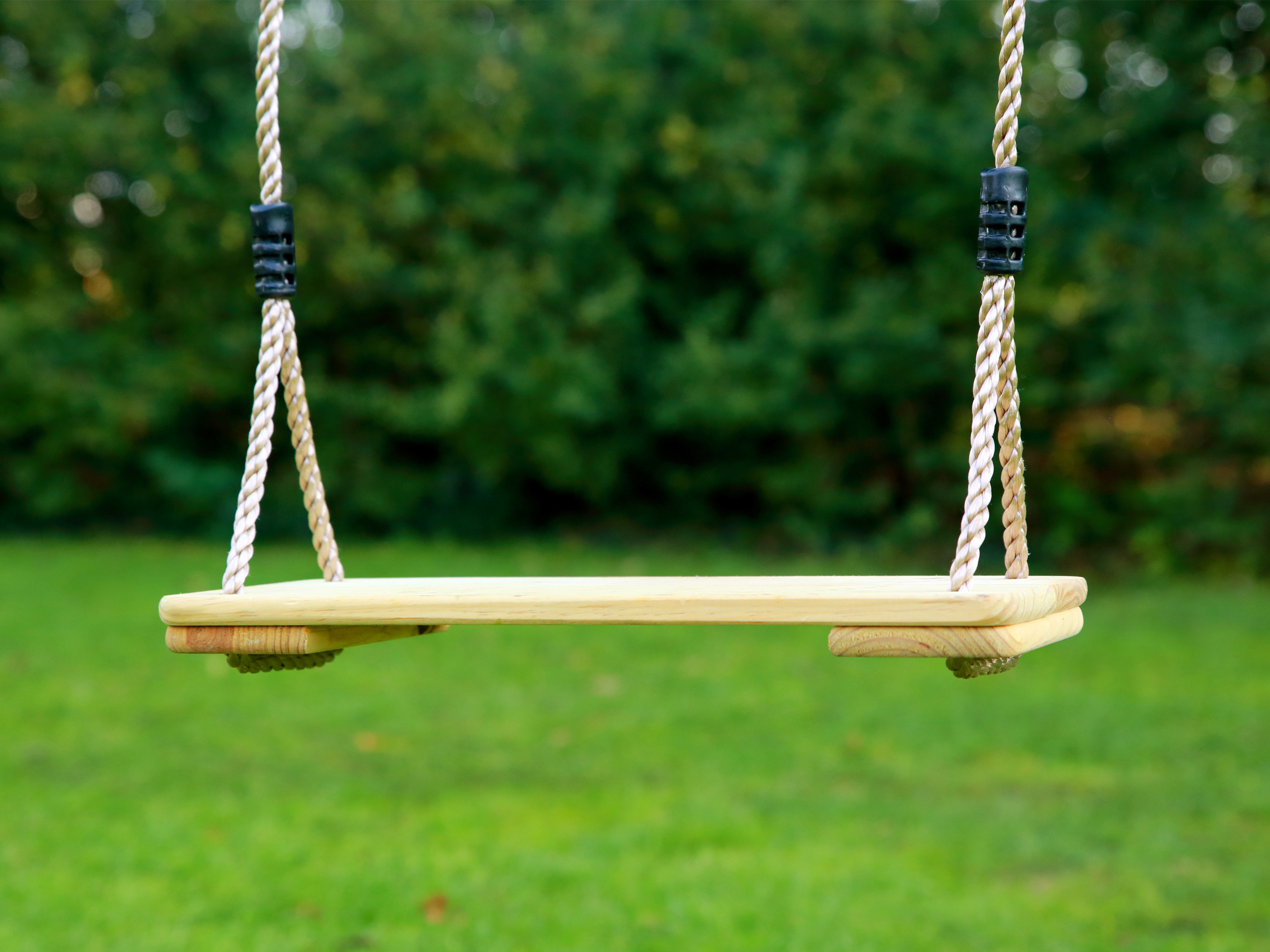 Holzschaukel mit Seil. Die Schaukel hängt in einem grünen, grasbewachsenen Bereich.