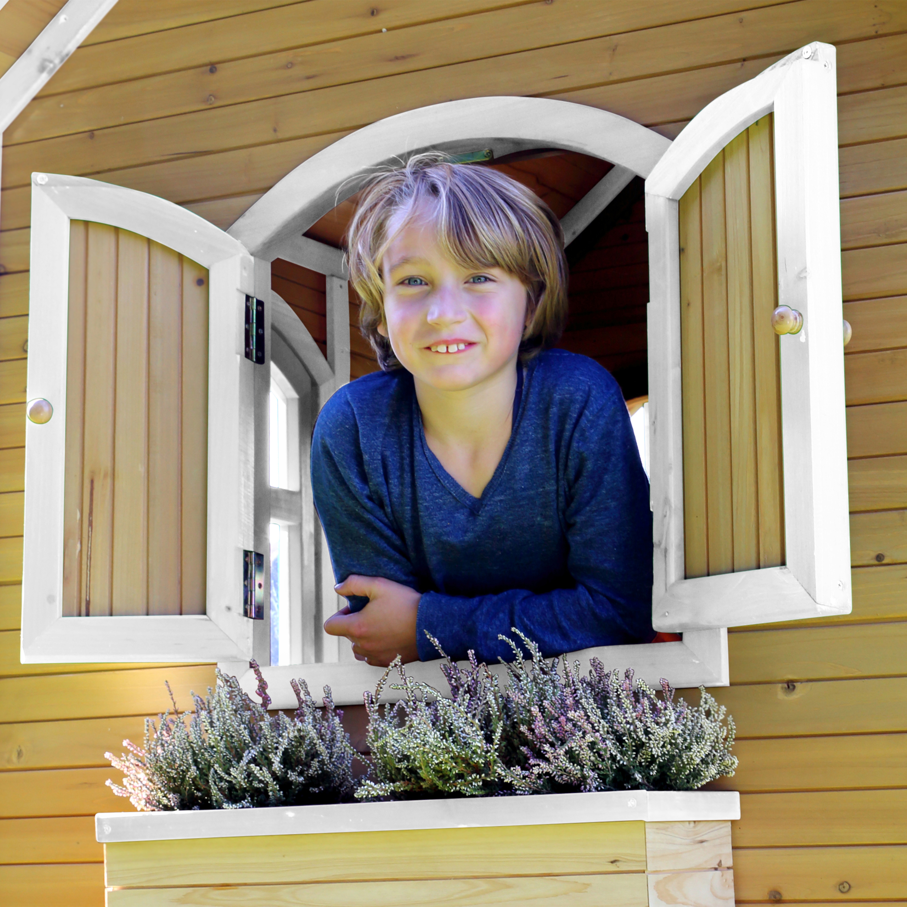 Ein Junge lächelt aus einem Holzfenster, mit Blumendekorationen im Fensterkasten.