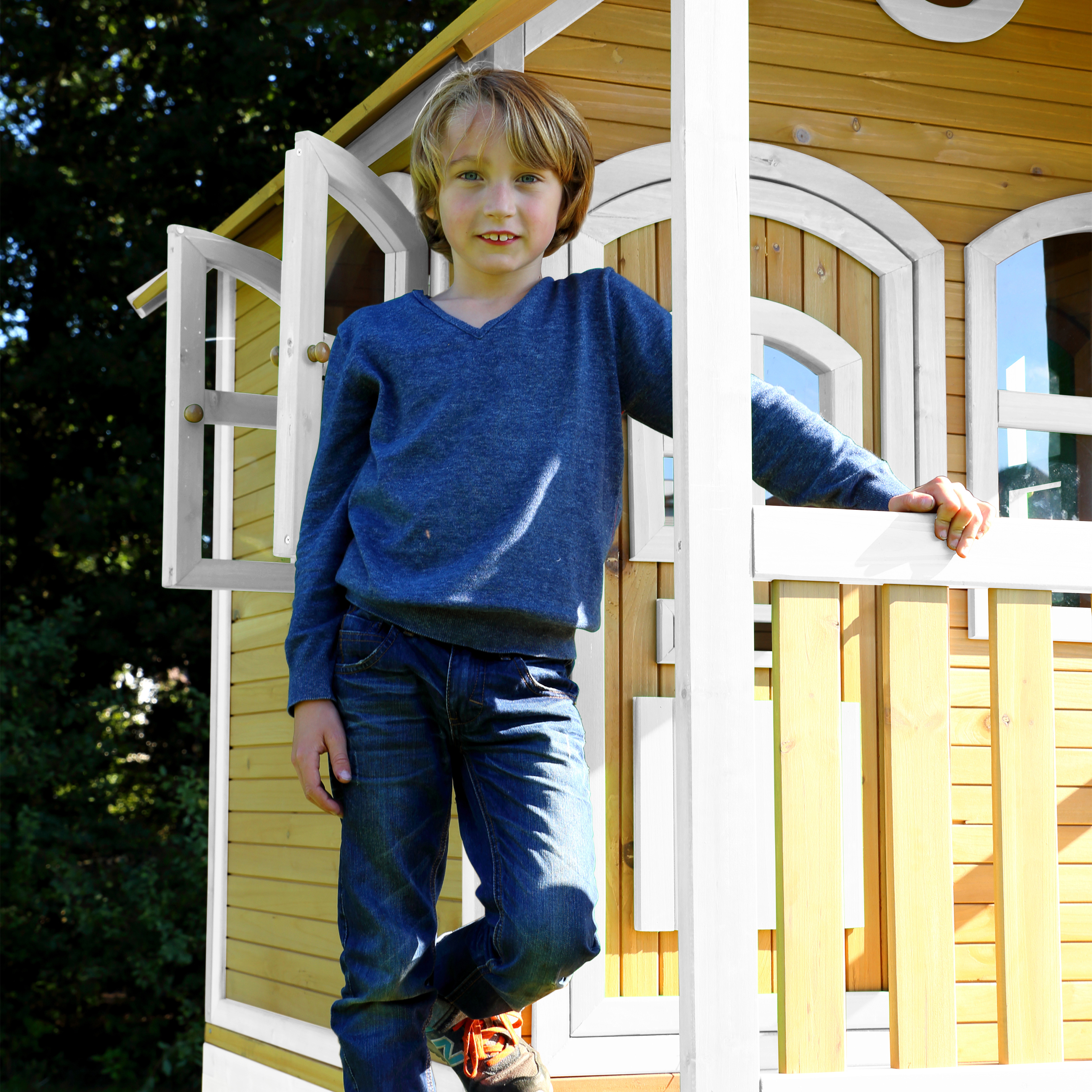 Ein junger Junge in einem blauen Pullover und Jeans steht in einem hölzernen Spielhaus und lächelt. Der Hintergrund sind grüne Bäume.