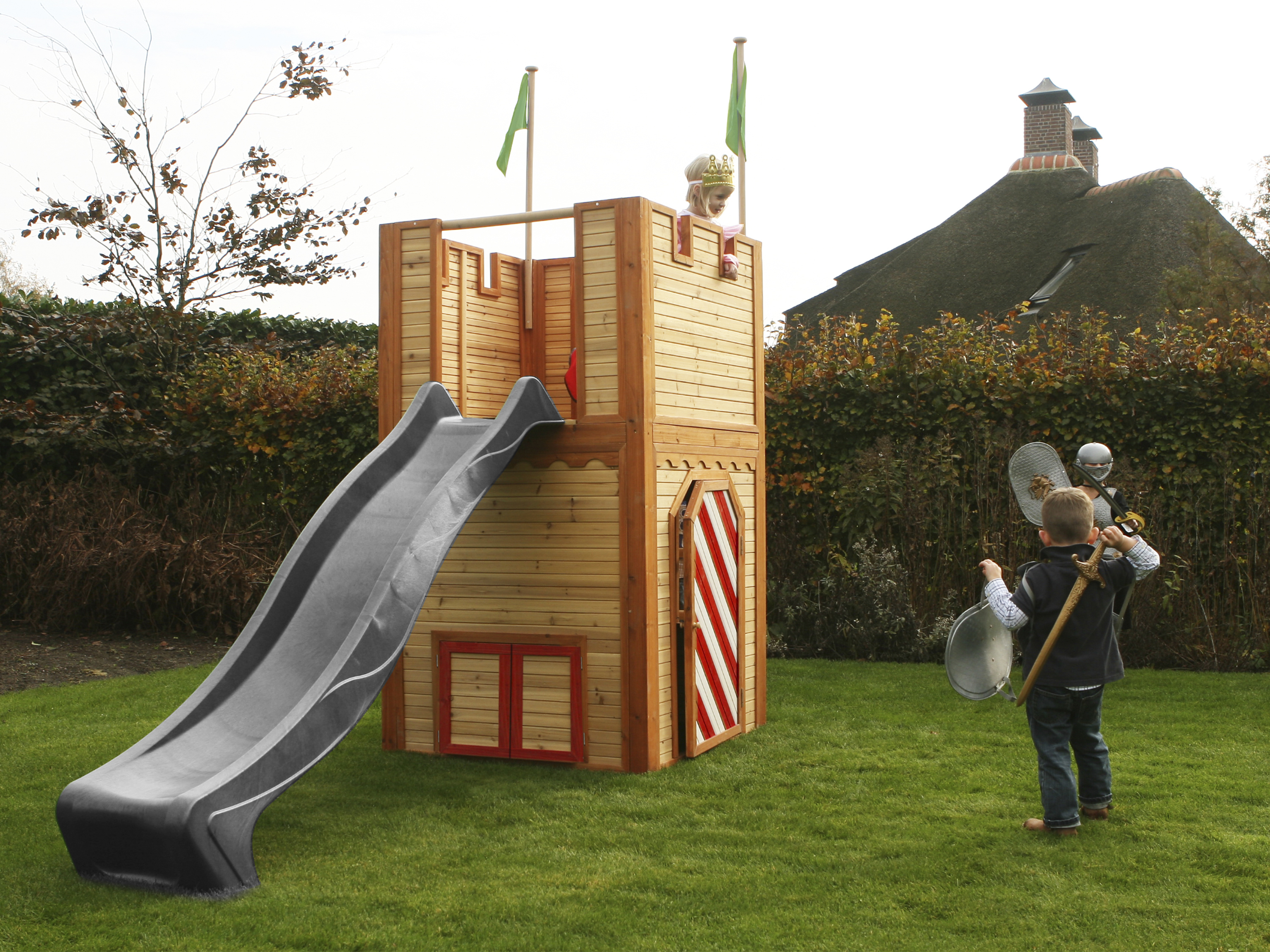 Ein Holzspielhaus mit Rutsche, und zwei Kinder spielen mit Schilden und einer Krone.