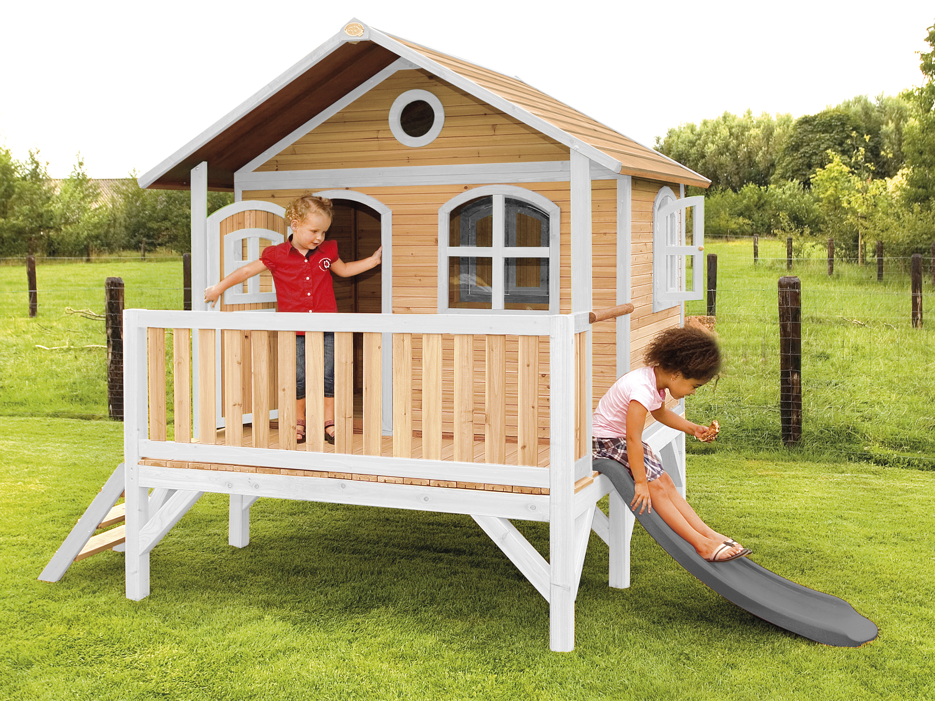 Kinder spielen in einem Holzspielhaus mit Rutsche. Grünes Gras und sonniger Tag.