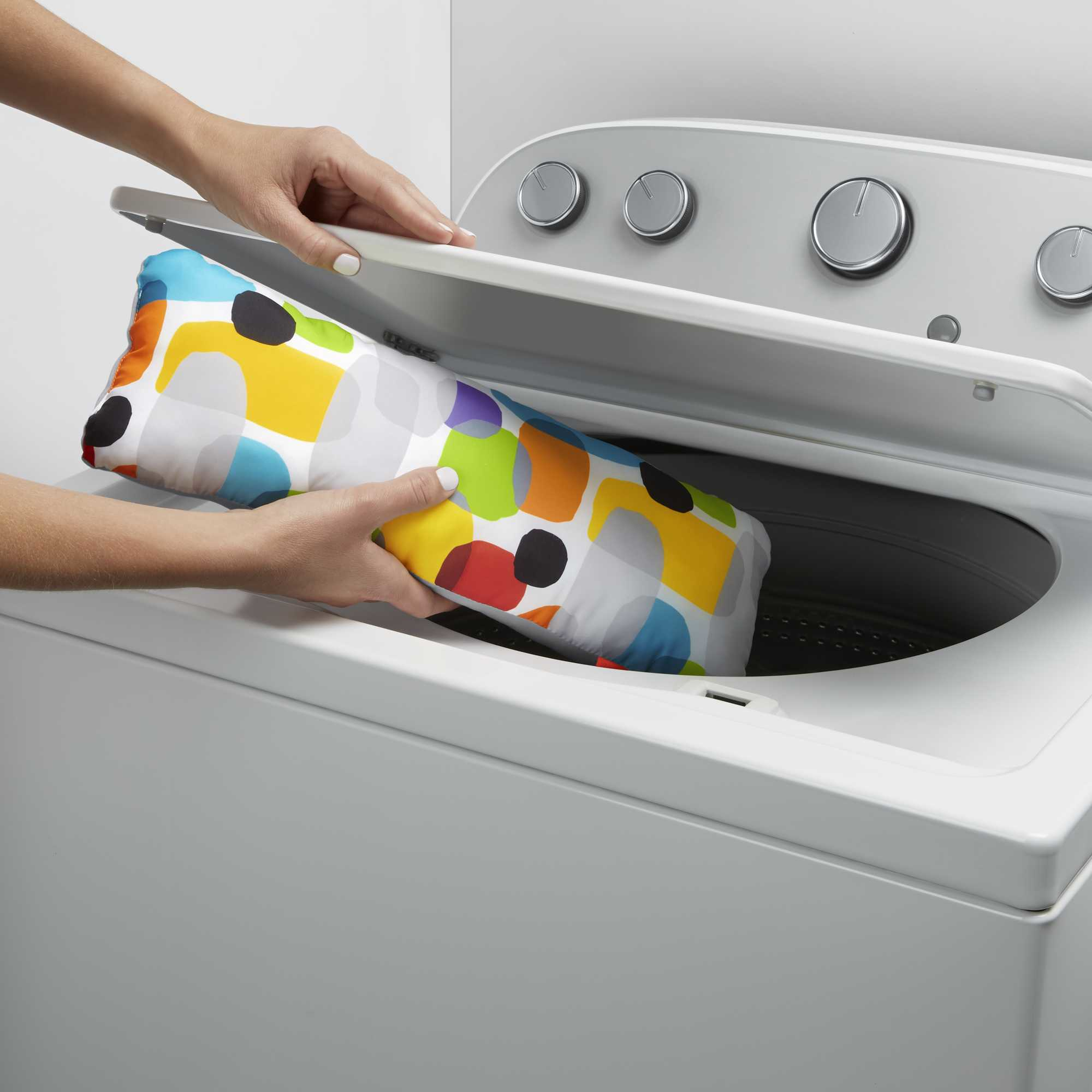 Hände legen ein buntes Kissen in eine Waschmaschine. Das Kissen hat ein Muster aus Formen.