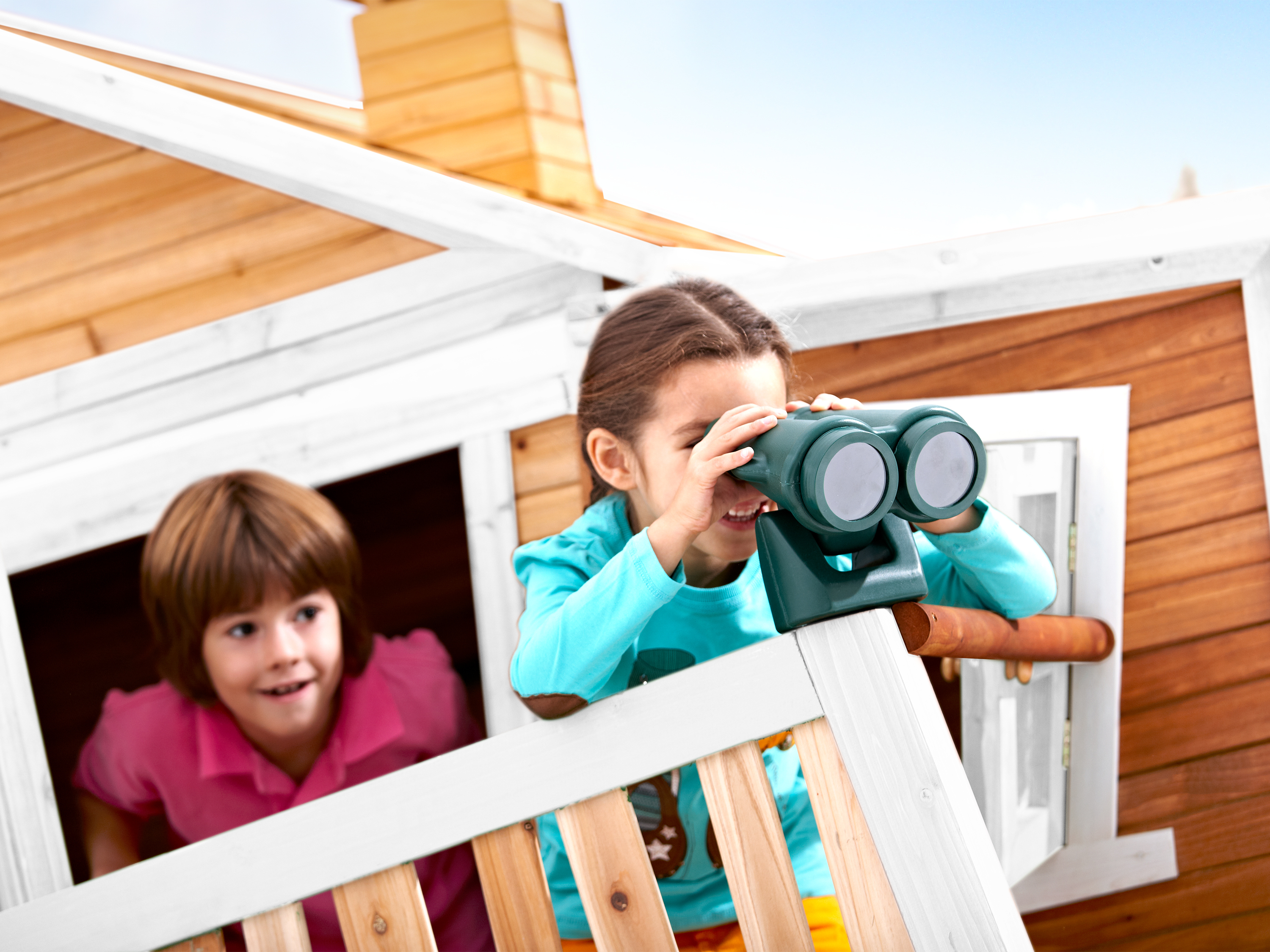 Zwei Kinder schauen durch ein Fernglas von einem Holzspielhaus aus. Das Haus ist braun und weiß.