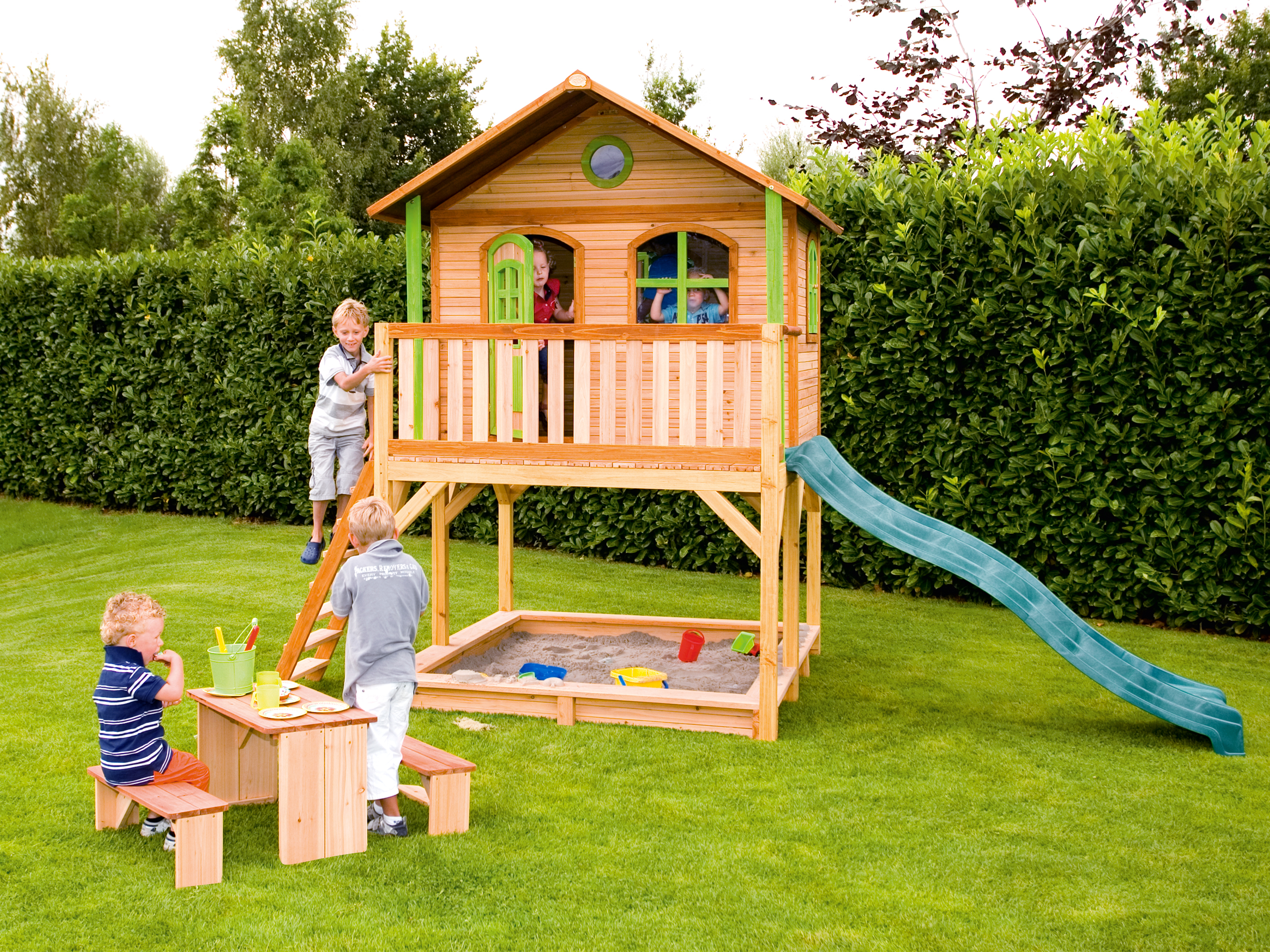 Kinder spielen auf einem Holzspielhaus mit Rutsche, Sandkasten und Tisch auf einer Wiese.