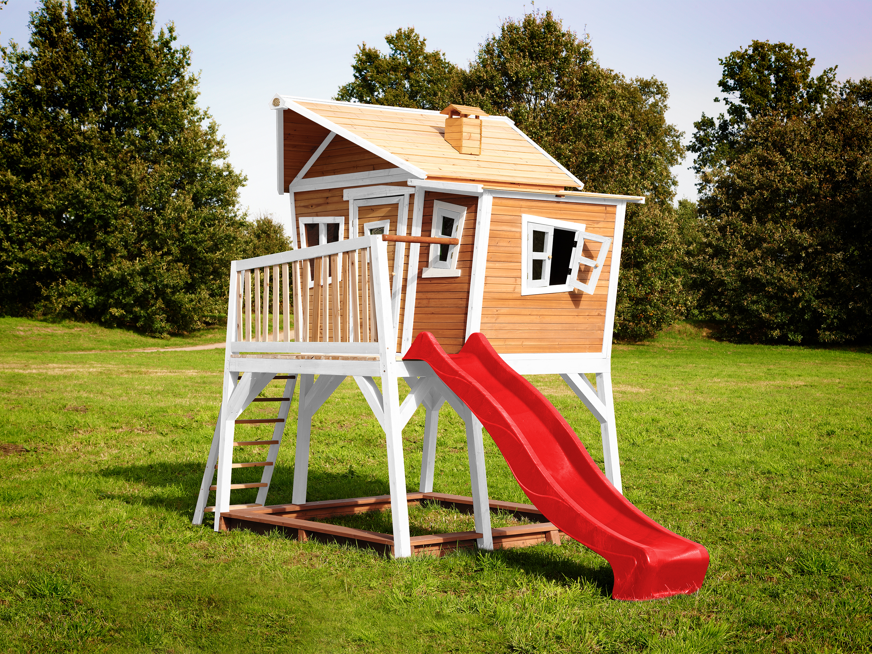 Ein Holzspielhaus auf Stelzen mit roter Rutsche auf einer Wiese unter klarem Himmel.